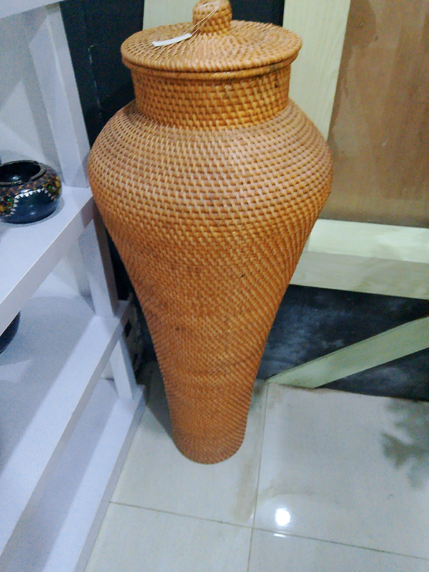 In Bunkate, Indonesia, a large, woven wicker pot with a lid stands on a tiled floor. The pot is light brown and has a textured pattern. To the left of the pot, a white shelf unit holds a small, dark, round decorative object with colorful markings. The background consists of a dark wall and a lighter wooden surface.