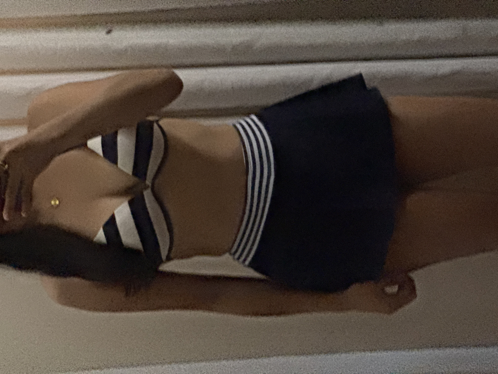 An individual is depicted standing indoors, reflected in a mirror or glass surface, against a background consisting of a light-colored curtain or textured wall on the right and a plain, light-colored wall on the left. The person wears a two-piece swimsuit. The top is a bikini bra with wide horizontal stripes of dark blue and white. The bottom is a dark blue skirted design featuring three narrow white horizontal stripes at the waistband. A gold pendant hangs from a necklace on the person's chest. The individual's left hand is partially visible near the top right of the frame, holding a device, with a gold ring on a finger. Long, dark hair is visible over the left shoulder. The lighting is low and indirect. The scene is located in Davao City, Philippines.