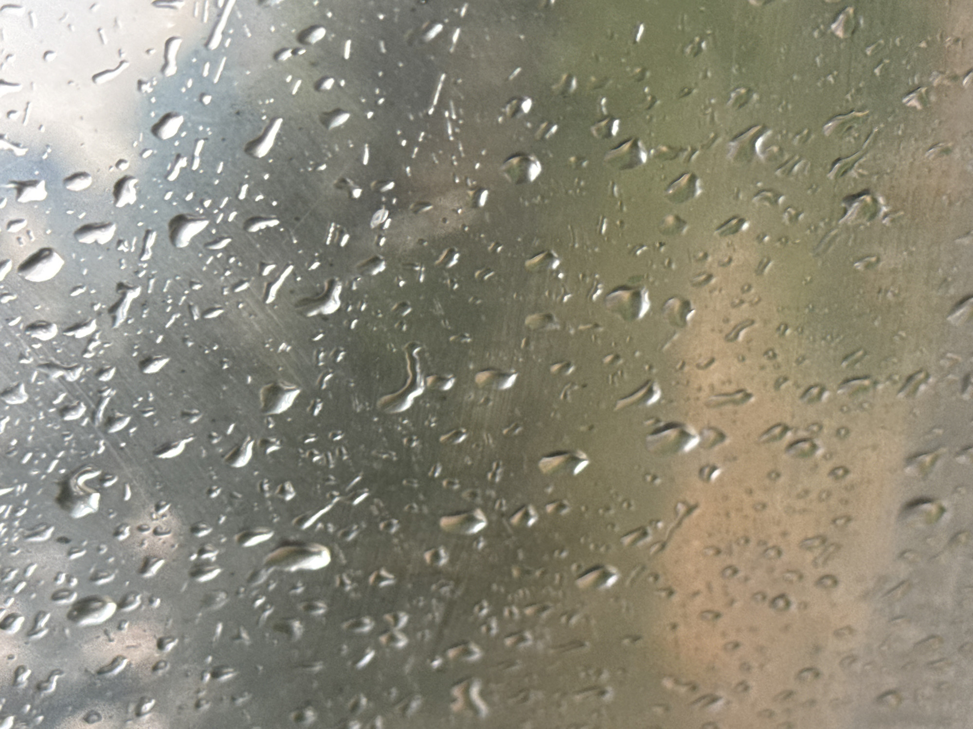 The image shows a close-up of a metallic surface covered in water droplets. The surface appears to be brushed metal, with visible streaks and a slightly reflective quality. The water droplets vary in size and shape, from small, round beads to larger, elongated streaks, indicating that they may have formed from rain or condensation. Some droplets are clear and sharp, while others are slightly blurred, suggesting variations in focus or the surface texture. The background is out of focus but suggests an outdoor environment, possibly a window or a metal structure. The lighting is diffuse, with some areas of brighter reflection on the water and metal. There are no discernible people, objects, or text in the image. The overall impression is one of a close-up, abstract study of water on a metallic surface, evoking a sense of natural elements interacting with man-made materials. The weather appears to be wet, likely raining or recently rained.