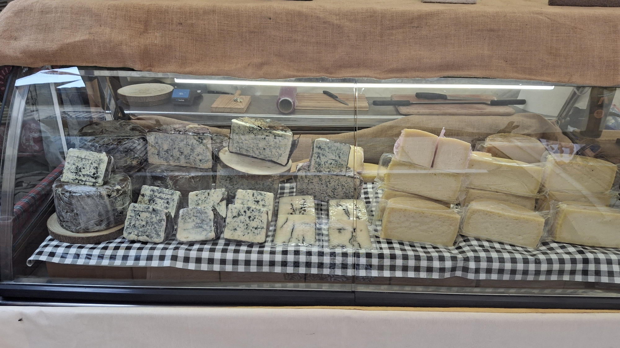 A cheese display case in an outdoor market in La Fresneda, Spain, showcases various types of cheese. The display case is made of glass and has a rustic burlap covering draped over the top. Inside, cheeses are arranged on a black and white checkered cloth.
In the foreground, several blocks of blue-veined cheese are cut into portions and individually wrapped in plastic. A larger, dark-rinded cheese is also visible, partially wrapped and resting on a wooden board. To the right, there are stacked blocks of pale yellow cheese, also individually wrapped.
In the background, more blue-veined cheeses are displayed, some on wooden stands. Behind the cheese, a roll of plastic wrap, a knife, and wooden cutting boards are visible, suggesting preparation or serving items. The overall impression is one of a well-organized and appealing artisanal cheese vendor at a local market.