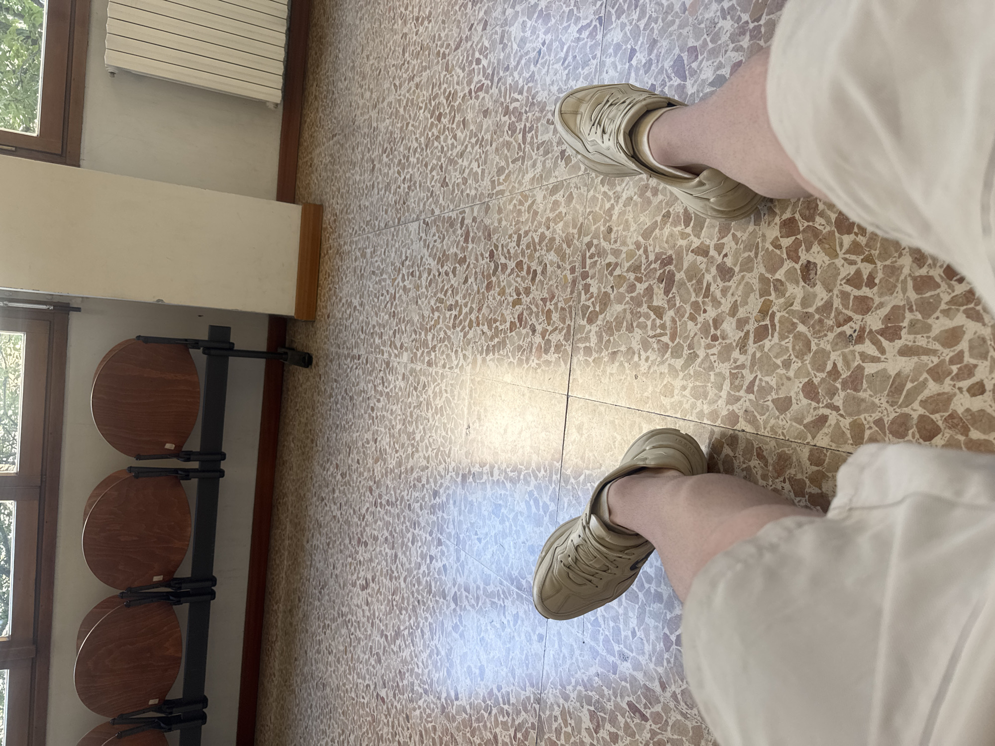 The image, taken from a first-person perspective looking down, captures the lower half of a person's body in an indoor setting, likely in Milan, Italy. The individual wears light-colored shorts and distinctive beige sneakers, with faint "GUCCI" branding visible on the side of one shoe. They appear to be standing or sitting on a light-toned, speckled terrazzo floor with visible grout lines. Bright natural light streams in from a nearby window, casting strong reflections and highlights across the polished floor tiles, suggesting it is daytime. To the left, a row of at least four circular wooden seats, attached to a black metal frame, indicates a public waiting area. Further left, a wooden-framed window looks out onto green foliage, while another window is partially visible below. Above the seating, a white horizontal fixture, possibly a radiator or blinds, is mounted on a cream-colored wall with dark wooden trim. The scene depicts a quiet moment in a public interior.