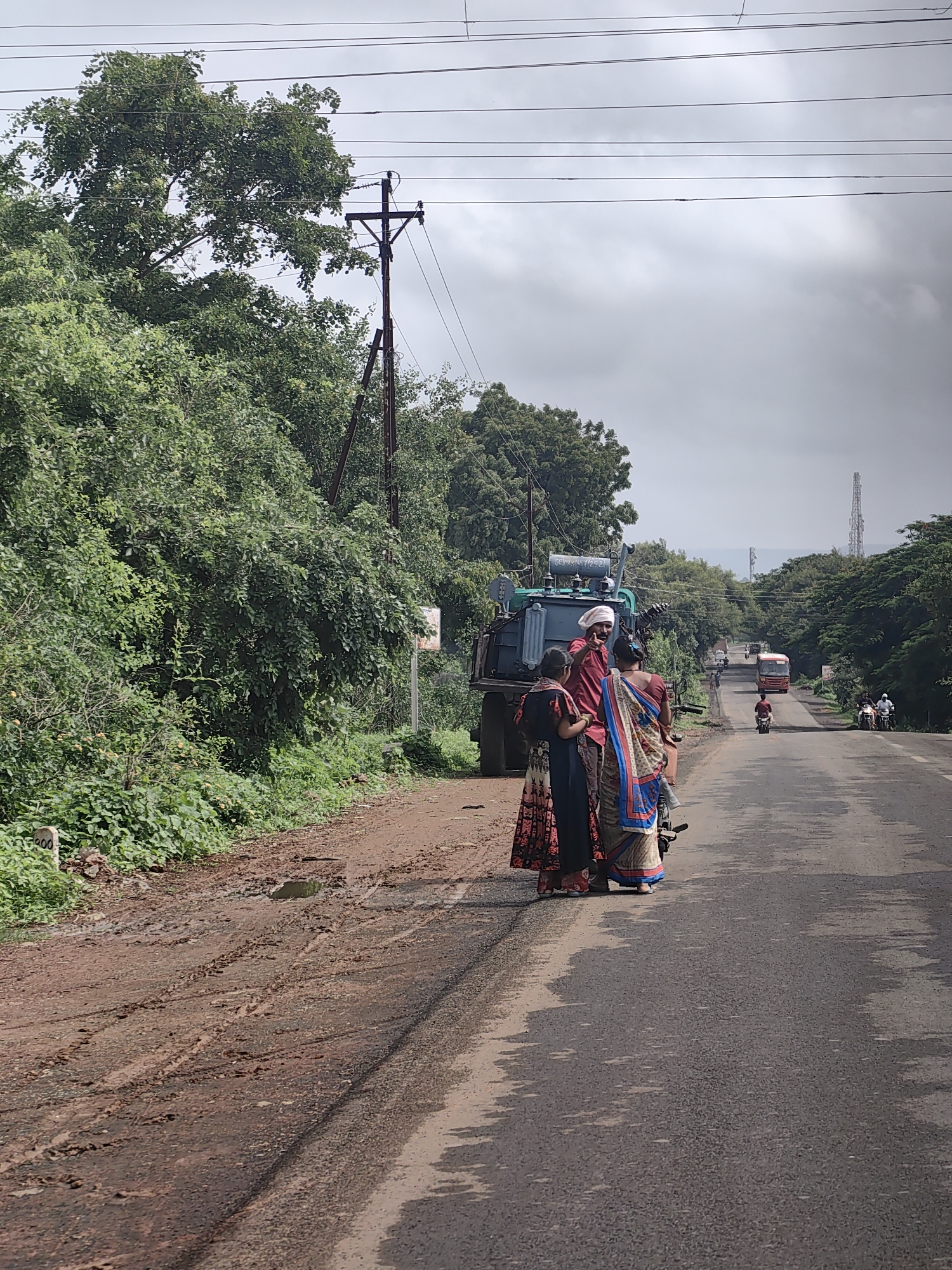A group of four people are gathered near a utility transformer on a dirt road, adjacent to a paved road in Kothli, India. The three individuals in the foreground, two women and one man, are dressed in traditional Indian attire. The man, wearing a white turban, points towards something off-camera while speaking to the women. The woman on the left wears a floral print dress, and the woman on the right wears a colorful saree. Behind them, a large green utility transformer is mounted on a vehicle. The scene is set against a backdrop of lush green trees and vegetation, with a cloudy sky overhead. In the distance, a bus and several motorcycles are visible on the paved road, indicating ongoing traffic. The dirt road is muddy and has tire tracks, suggesting recent rain or wet conditions. A power line pole with wires is also visible to the left of the scene.