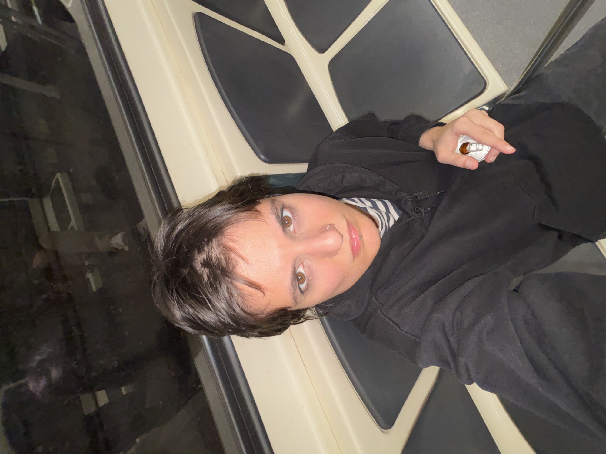 A person with brown hair and eyes, wearing a black hoodie and a striped shirt, is seated on a train. The person is holding an open can in their right hand. The train car features cream-colored panels with a black pattern. The scene appears to be inside a metro train in Sofia, Bulgaria, based on the distinctive interior design of the train car.
