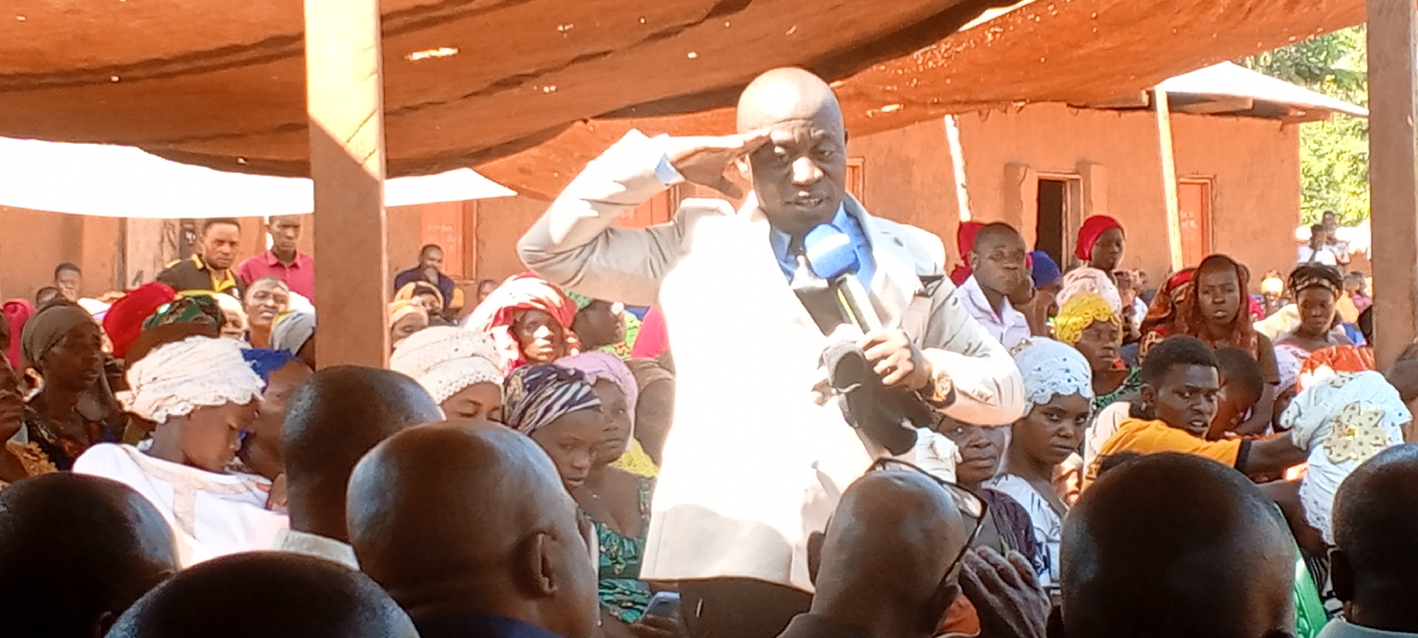 The image shows a man speaking into a microphone in front of a crowd. He is wearing a light-colored suit and has his right hand raised to his forehead in what appears to be a salute. The crowd consists of men, women, and children, many of whom are wearing head coverings. The setting appears to be outdoors, with several buildings visible in the background. Based on the prompt, the location is Kalemie, Democratic Republic of the Congo.