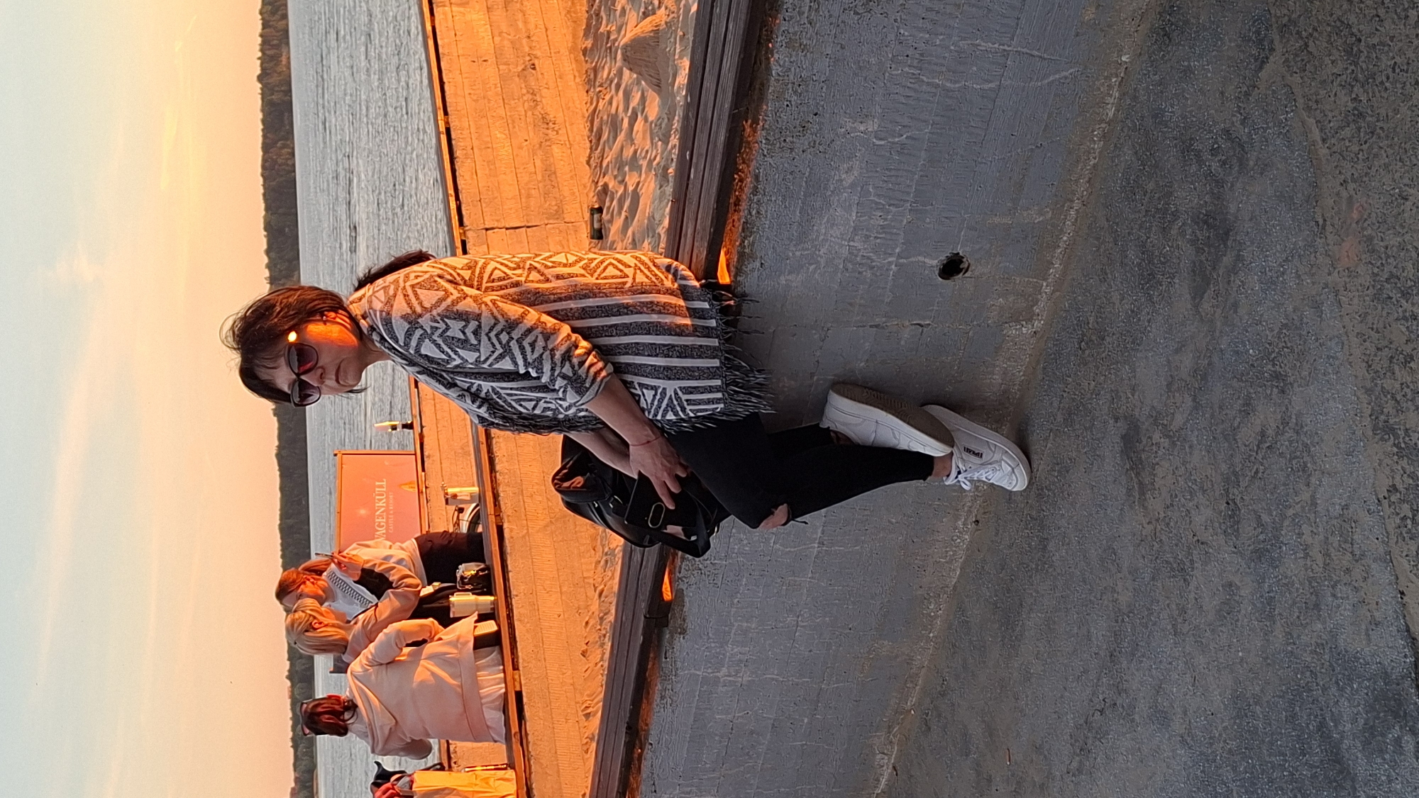 A woman in her 40s or 50s with dark hair and sunglasses is sitting on a concrete ledge at sunset. She is wearing a patterned poncho over a black top and ripped black jeans, with white sneakers. She is holding a mobile phone and has a black bag next to her. In the background, two other women are visible, one taking a photo with her phone. They are standing near a sign that says "VÄGENKÜLL". The scene appears to be a waterfront area, possibly a beach or promenade, with sand and a body of water visible. The lighting suggests it is late afternoon or early evening. This location is likely in Tallinn, Estonia, given the visible sign and the context of the other individuals.