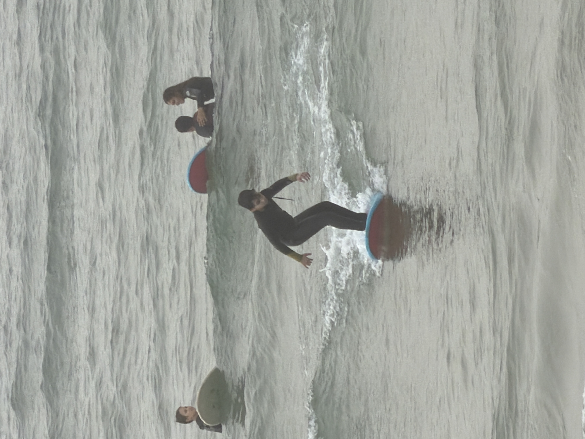 The vertically oriented image captures a water sports scene featuring four individuals in dark wetsuits engaged in bodyboarding or small-board surfing, set against a backdrop of rippling ocean water. The context suggests the location is Tel-Aviv, Israel.
In the upper portion of the frame, two individuals are visible. One, lying prone on a dark bodyboard, is partially obscured. The other is positioned next to a red and blue bodyboard. Towards the middle of the image, a third person is actively riding a wave on a blue and red bodyboard or small surfboard, their arms outstretched for balance amidst the white, foamy water. In the lower left area, the head and upper torso of a fourth individual are partially visible beside a light-colored bodyboard.
The water's surface exhibits a pattern of small waves and foam, indicating active but not rough conditions. The scene is illuminated by daylight, with soft or no discernible shadows, possibly suggesting an overcast day. No text is visible in the media file.
