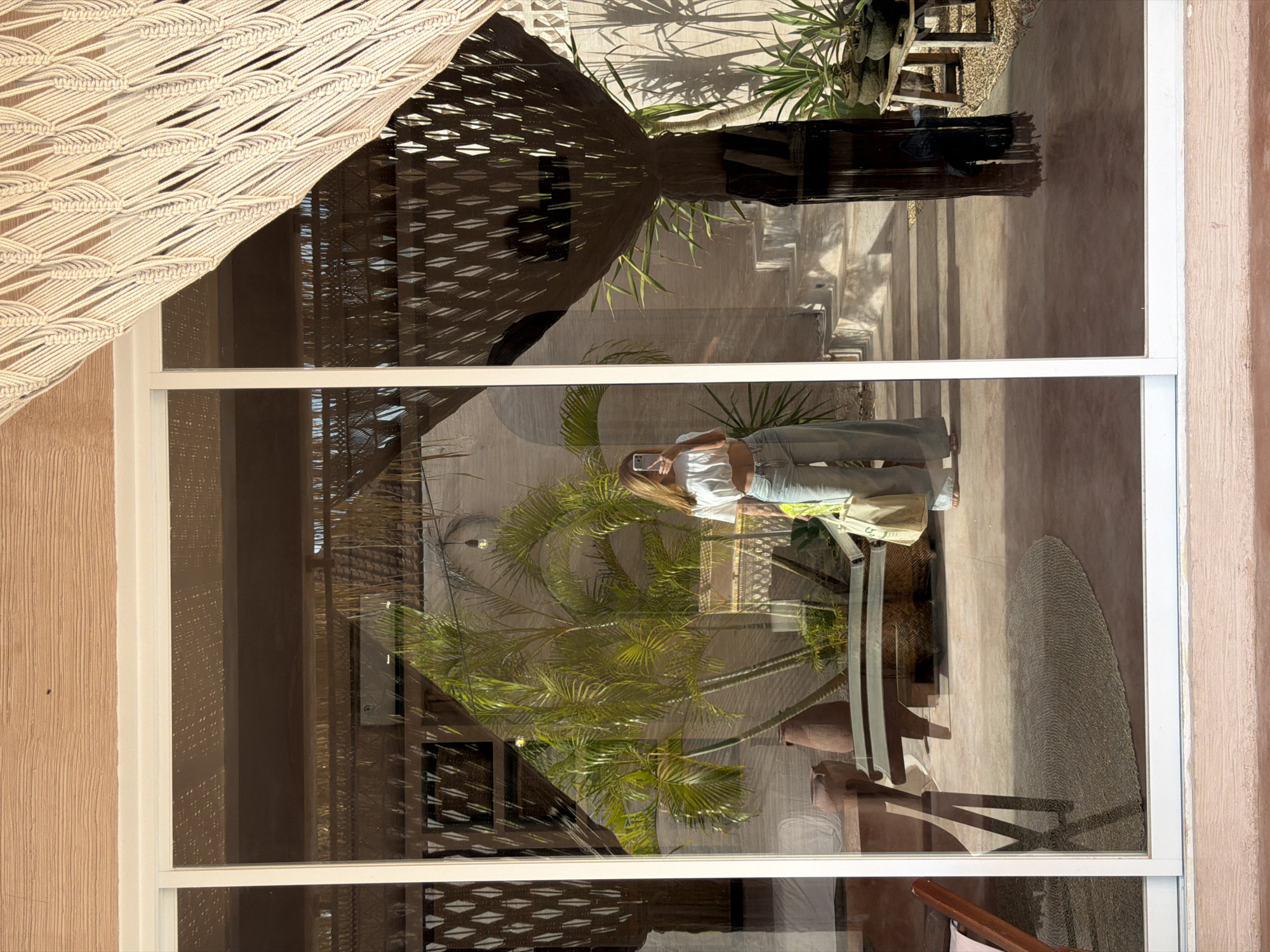The image shows a person reflected in a window in North Kuta, Indonesia. The person is holding a phone and wearing jeans and a white top. The reflection shows a courtyard with palm trees, furniture, and a macrame shade. The building exterior is a light peach color.