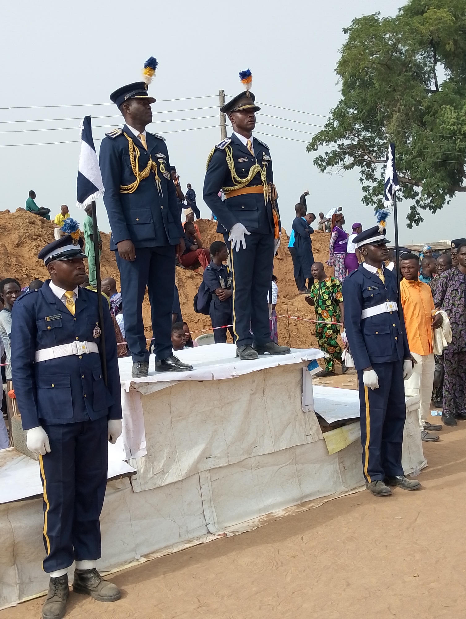 The image depicts an outdoor formal event or ceremony in Kubwa, Nigeria, featuring four men in striking dark blue military-style uniforms. The uniforms are adorned with gold trim, white gloves, and peaked caps topped with a distinctive blue and yellow plume. Two of the uniformed men stand prominently on an elevated, white fabric-covered platform, wearing elaborate gold cord lanyards (aiguillettes) across their chests. A third uniformed individual stands on a lower section of the same platform, also displaying gold uniform details and a similar cap plume. The fourth uniformed man stands on the bare dirt ground in the foreground, distinguished by a white belt and gold stripe on his trousers. All four maintain a solemn, attentive posture, gazing forward or slightly to the right.
In the background, a crowd of spectators is gathered, with some individuals sitting casually on a large dirt mound and others standing. Two black and white flags are partially visible, positioned behind the uniformed personnel. The setting indicates a daytime event under an overcast or hazy sky, contributing to diffused lighting without harsh shadows. Cues to the location include the expansive dirt ground, the presence of utility poles and wires against the sky, and a large tree with green foliage on the right side of the frame, suggesting a public or open area.
Visible text on the name tag of the uniformed man standing on the ground reads "OLUWAJUWON". Additionally, the name "D. A. OSAMA" is discernible on the name tag of the uniformed man on the lower platform section to the left.