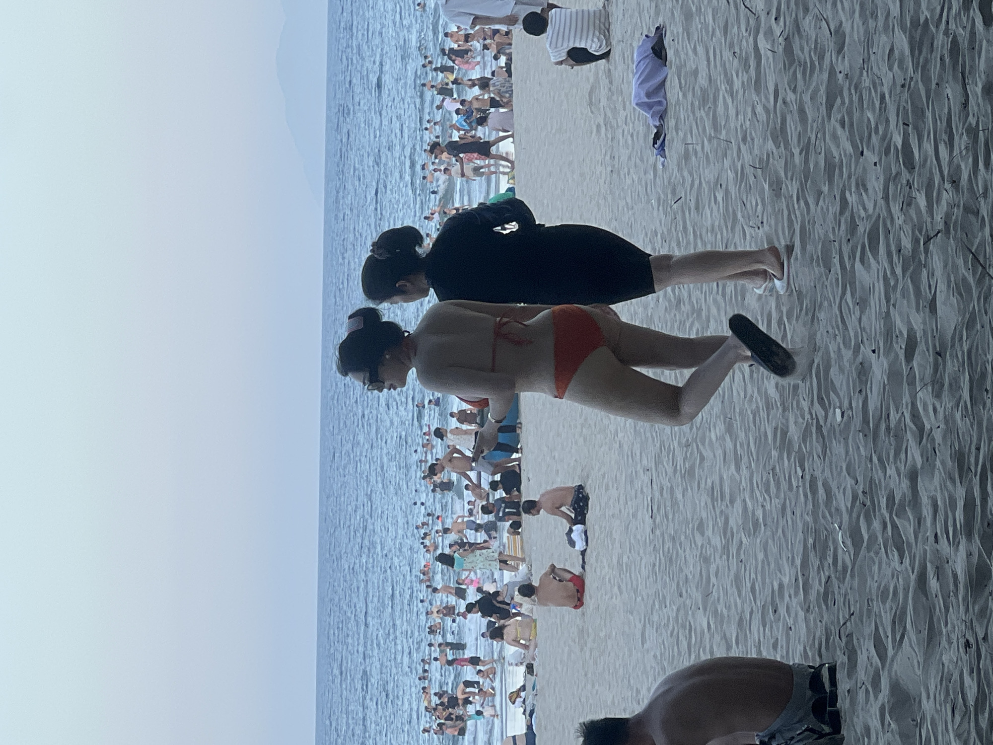 The image shows a crowded beach with many people in the water and on the sand. The scene appears to be at a popular tourist destination in Da Nang, Vietnam, likely My Khe Beach, known for its long stretch of white sand and clear blue waters. In the foreground, two women are standing on the sand, facing away from the camera. One is wearing an orange bikini, and the other is wearing a black swimsuit. Behind them, a large number of people are swimming, playing in the water, or relaxing on the beach. The water is calm with gentle waves, and the sky is a pale blue, suggesting it might be late afternoon or early morning. In the distance, the outline of mountains can be seen against the horizon. The overall atmosphere is one of a lively beach scene on a warm day.