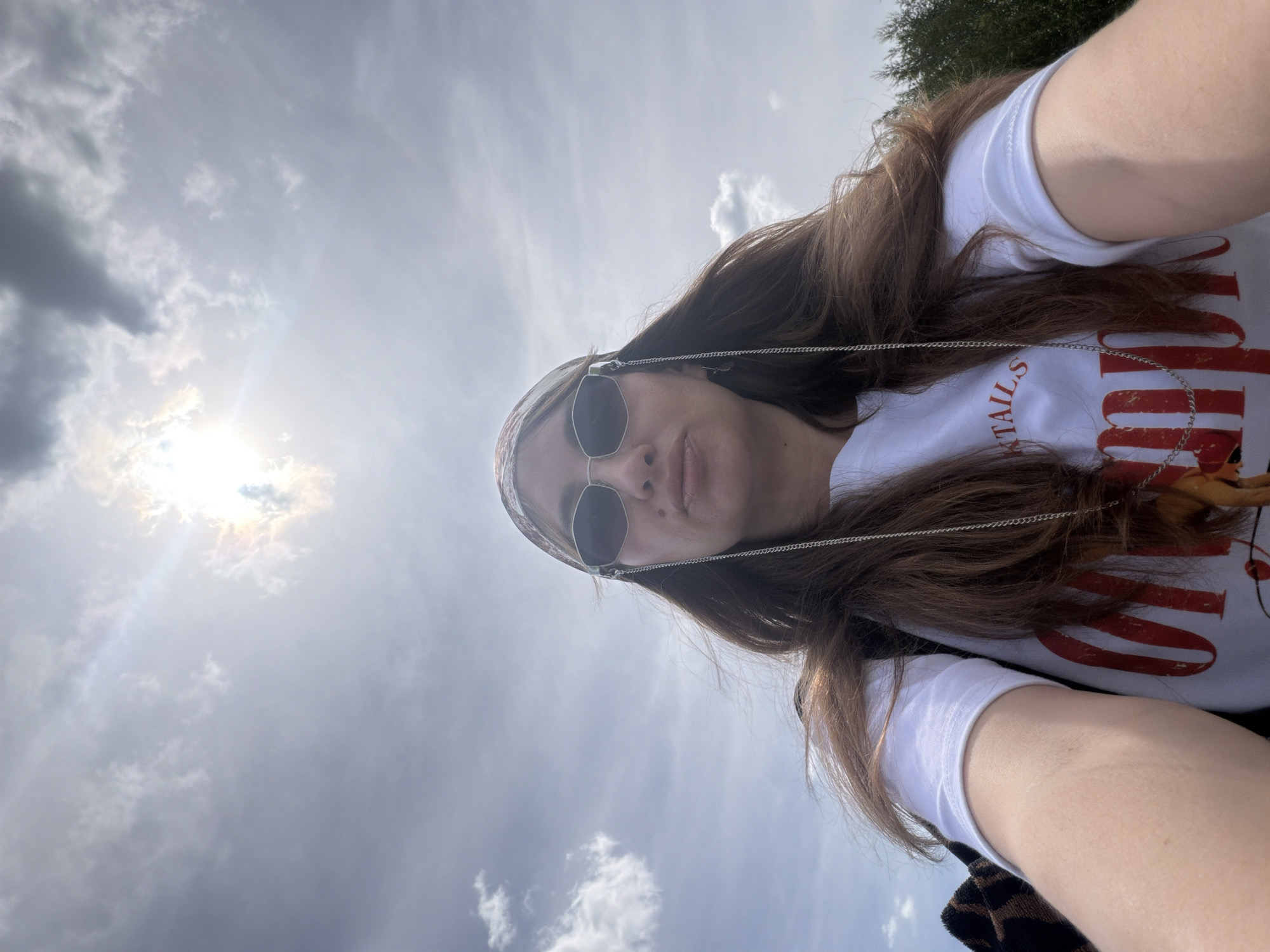 An outdoor, waist-up, selfie-style photograph shows a woman with long brown hair wearing a white t-shirt with red lettering that reads "COCKTAILS", a patterned bandana, and sunglasses with a silver chain. The background is a bright, cloudy sky with the sun partially obscured by clouds. Trees are visible in the lower right corner. The woman is looking at the camera with her lips pursed.