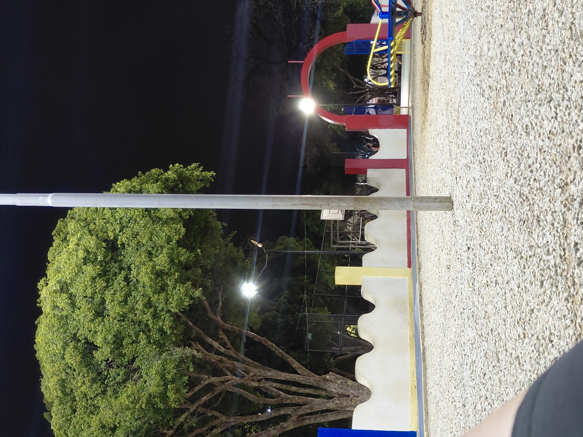 The image depicts a nighttime scene of a public park or plaza in Santa Ana, Venezuela. The sky above is uniformly dark black, indicative of night, with distinct lens flare artifacts originating from the several bright artificial light sources within the park.
In the left and background, large, mature trees with dense, vibrant green foliage are prominently featured, brightly illuminated by the park's lighting. The sturdy, multi-branched trunk of a significant tree is visible on the far left.
A tall, light-grey utility pole stands vertically in the left foreground, partially obscuring the tree and sky. Across the midground, a low, undulating white wall, stylized with wave-like peaks and troughs, extends horizontally. Sections of this wall are accented with bold colors: blue on the far left, yellow in the middle-left, and red towards the right. Behind the wall on the left, a white basketball backboard and hoop are partially visible under the glow of a streetlight.
On the right side of the frame, a distinct red arched gateway, featuring vertical blue bars, marks an entrance or focal point within the park. Adjacent to this archway, brightly colored yellow and blue components of what appears to be playground equipment, possibly a merry-go-round, are visible.
The entire foreground and much of the visible ground surface consist of a uniform layer of small, irregularly shaped, light-colored (off-white to beige) gravel or crushed stone. A dark, curved shape, consistent with the edge of a human limb or article of clothing, is present in the extreme bottom-left corner of the frame. No identifiable individuals or discernible actions are visible within the depicted scene.