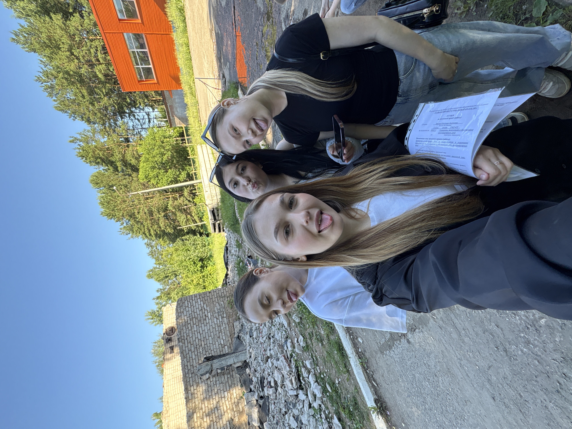 Four young women are taking a selfie outdoors. The woman in the foreground is wearing a black jacket over a white shirt, and has long blonde hair. She is holding a document. The woman to her right is wearing a black t-shirt and blue jeans, with blonde hair tied in a ponytail. She is holding a black mobile phone. To the left of the foreground woman is a woman in a white t-shirt with dark hair pulled back. Behind her, and slightly to the right, is a woman with dark hair wearing a white top and sunglasses on her head. The background shows a ruined brick building on the left, with piles of rubble. Further back, there are trees and an orange building with windows. The ground is paved with dirt and gravel. The sky is blue with no clouds. The scene appears to be an abandoned industrial or construction site. The location is Chaikovsky, Russia.