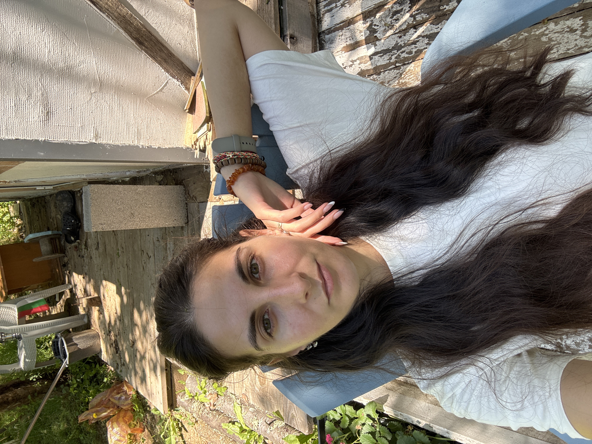 A woman with long, dark hair is seated outdoors, resting her arm behind her head. She is wearing a white t-shirt and several bracelets on her left wrist. She is situated on a blue chair, with a weathered wooden deck beneath her. The background reveals a rustic, outdoor setting with vegetation, construction materials, and a partially visible structure that appears to be a small building or shed. The scene suggests a relaxed, possibly rural, environment. The provided location is Dramsha, Bulgaria.