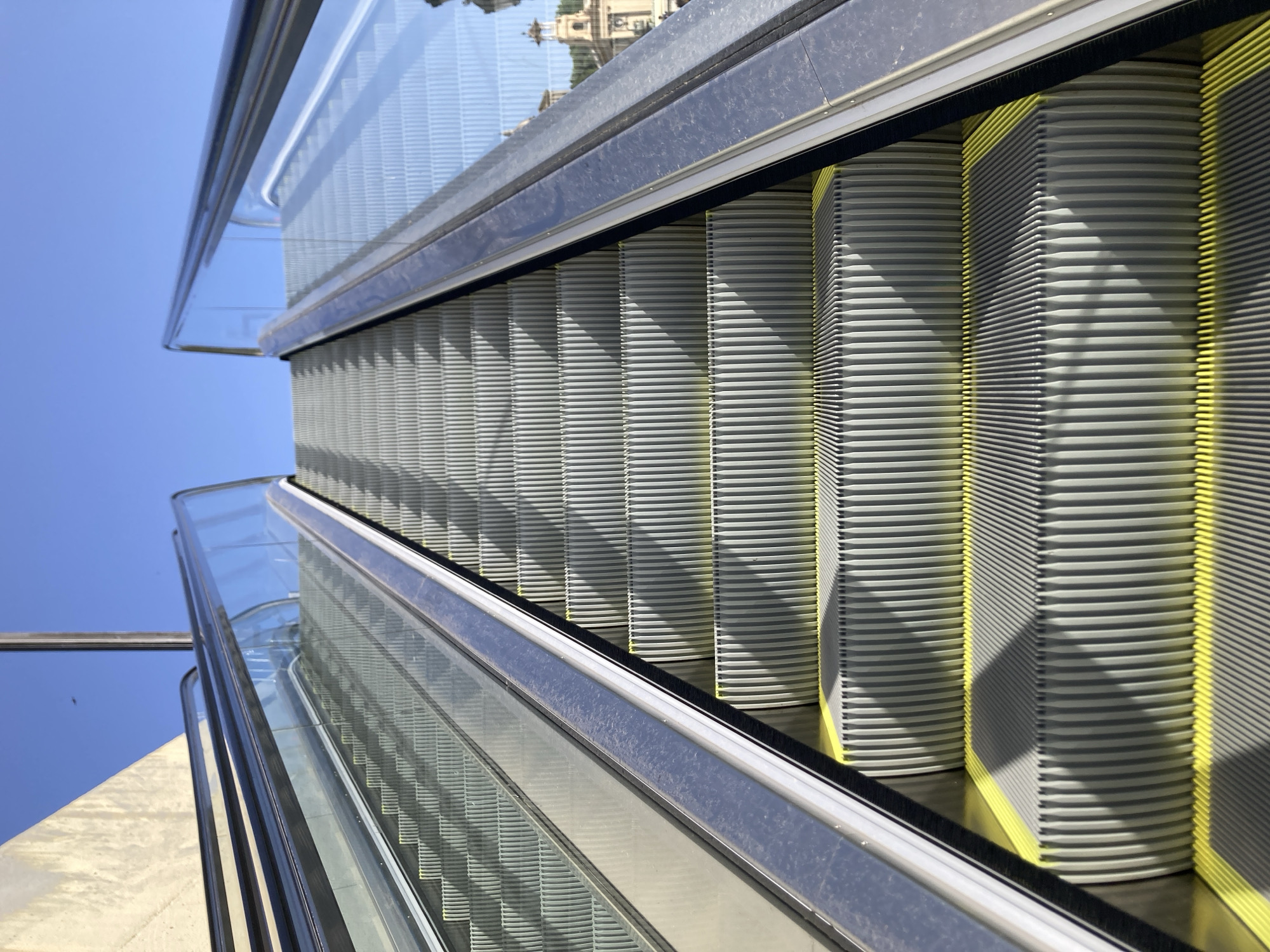 This image shows a close-up perspective of an outdoor escalator in Barcelona, Spain, ascending towards a clear blue sky. The escalator's metal steps are ribbed and a bright yellow strip runs along the edge of each step. Glass railings with metal handrails line both sides of the escalator. To the left, a textured concrete wall is visible, likely part of a building. The right side of the image shows a blurred background of buildings, including a church with a dome and towers, suggesting an urban environment. The lighting indicates it is daytime, with strong shadows cast across the escalator steps. There are no people visible on the escalator.