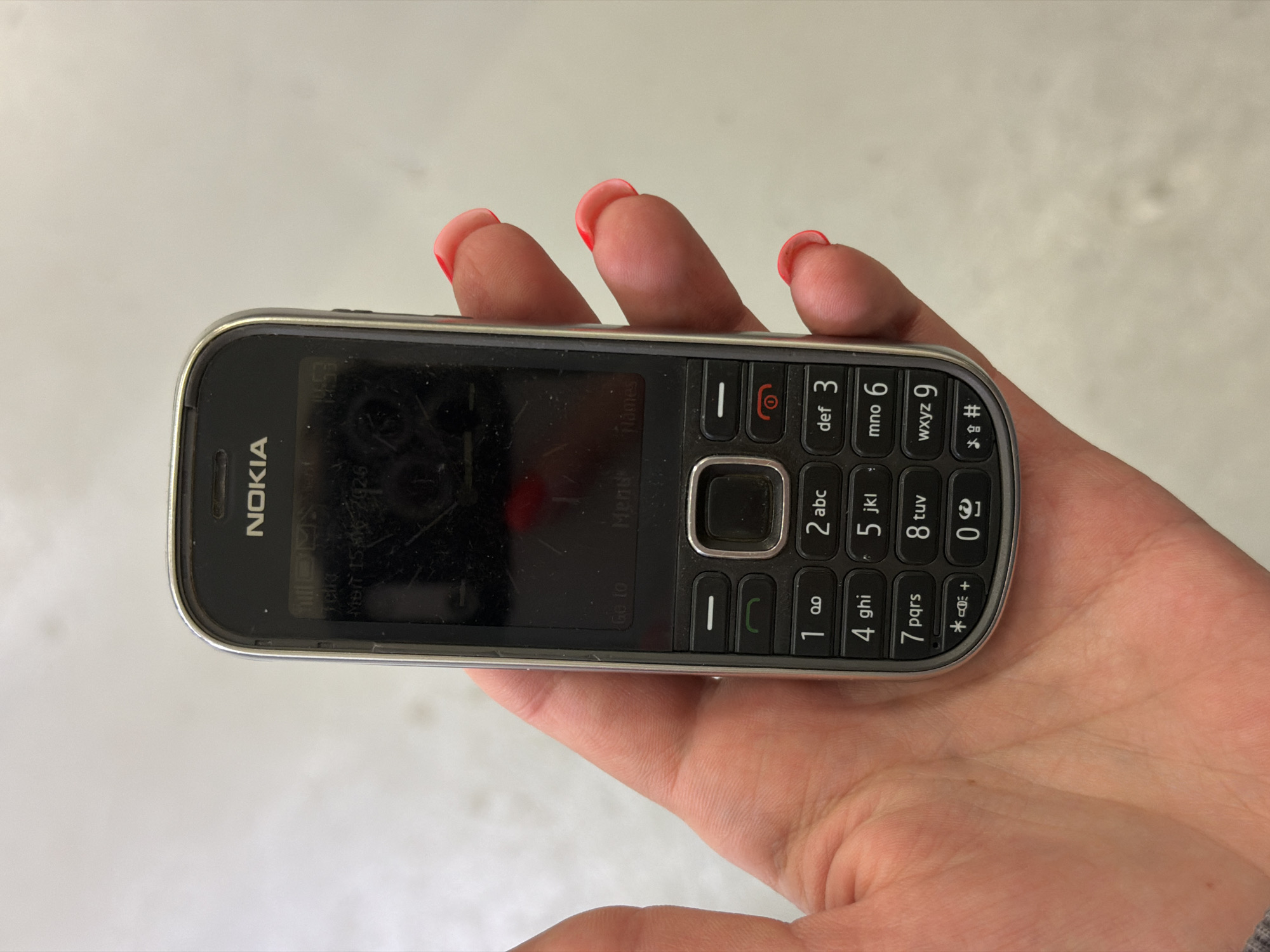 An individual's left hand, with bright pink nail polish, is holding a black Nokia feature phone. The phone screen displays "NOKIA" at the top, followed by the date and time "Mon 15-Aug-2026 10:53". Below this, there are menu options. The keypad has numerical buttons with corresponding letters and symbols. The background is a blurry, light-colored surface, possibly a floor or wall, suggesting an indoor setting. The scene is not identifiable as being in Viimsi, Estonia.