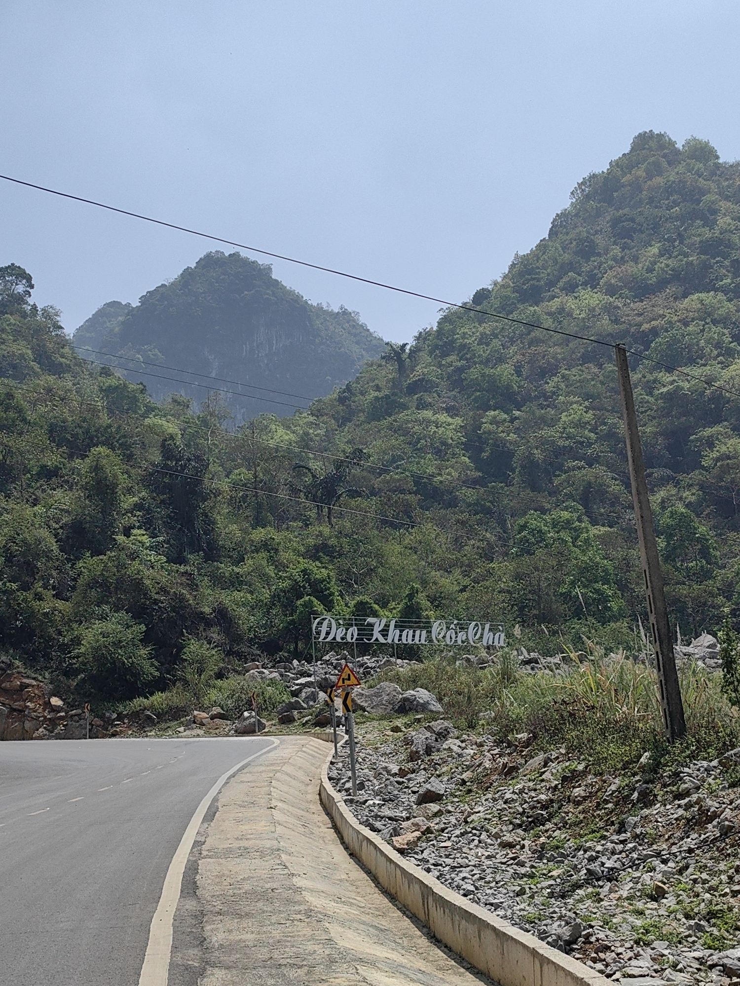 The image shows a road curving through a mountainous, heavily vegetated landscape. A sign reading "Đèo Khau Cốc Chà" is visible on the right side of the road. The road has a concrete edge. A utility pole stands near the sign. The mountains in the background are covered in dense foliage. The sky is overcast. The location is likely in the vicinity of Xã Xuân Trường, Vietnam, given the sign.