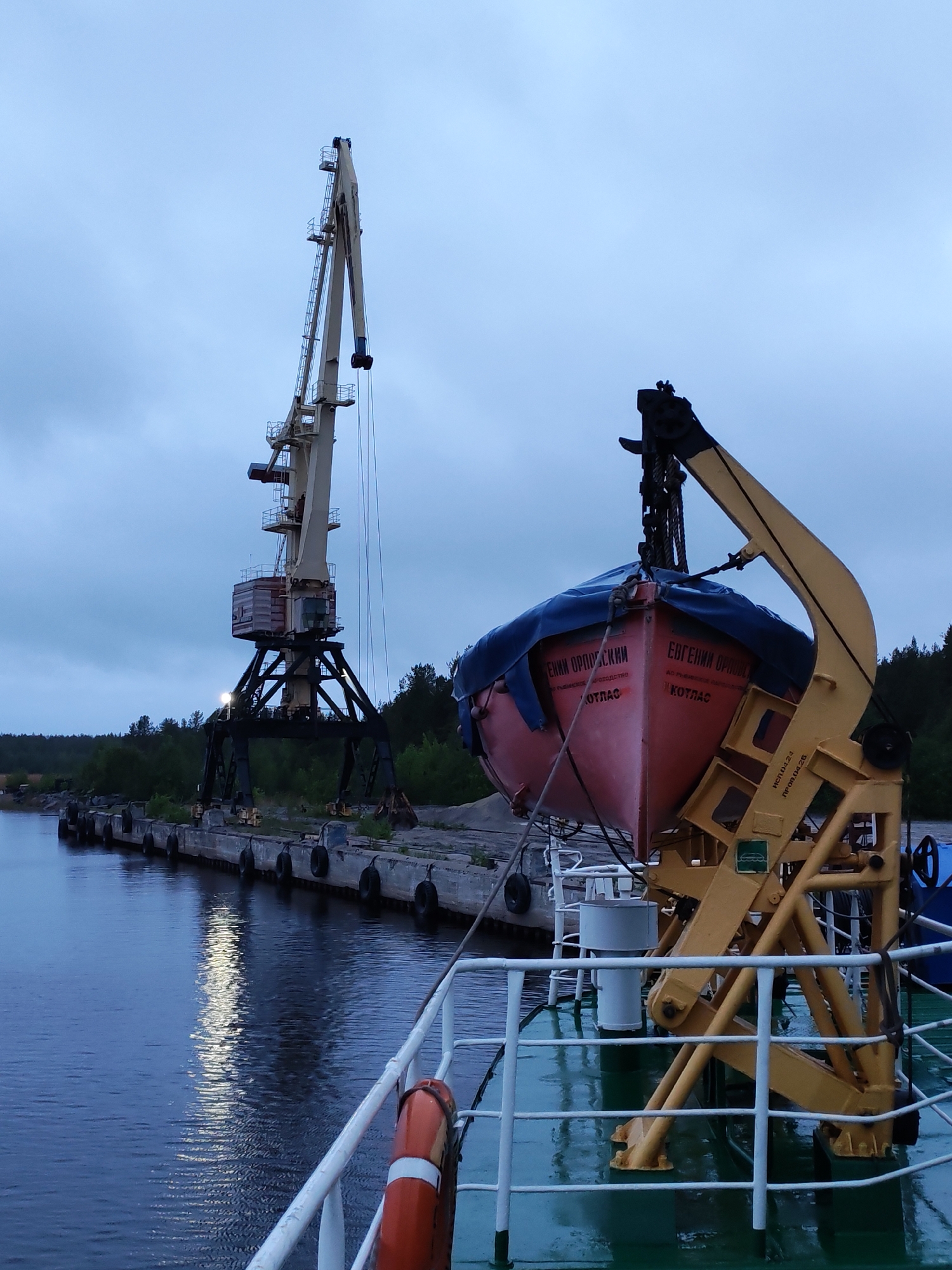 The image shows a scene at a port or dockside, likely in Shalsky, Russia based on the signage on the lifeboat. A large, stationary crane stands on the dock, with a lifeboat suspended in the air by a smaller, yellow-painted crane on a vessel in the foreground. The dock is concrete with rubber tires used as bumpers along the edge facing the water. The water reflects the light from the crane and possibly other sources, indicating a possibly dusk or dawn scene. The sky is overcast. The lifeboat has Cyrillic lettering on its side. The vessel with the yellow crane has a white railing and a green deck. An orange life preserver is attached to the railing. Trees are visible in the background.