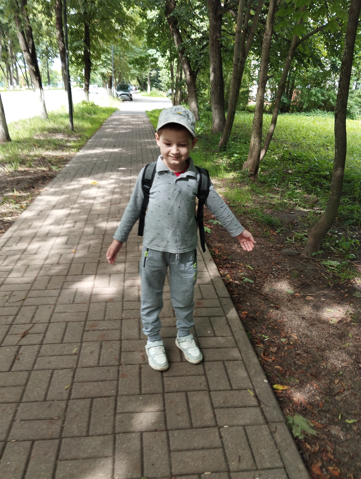 A young boy wearing a grey polo shirt, grey sweatpants, a grey baseball cap, and white sneakers stands on a paved walkway lined with trees. He has a black backpack on. The walkway is bordered by grass and trees on the right and a road with cars and more trees on the left. The scene appears to be in a park or along a tree-lined street in Kingisepp, Russia.