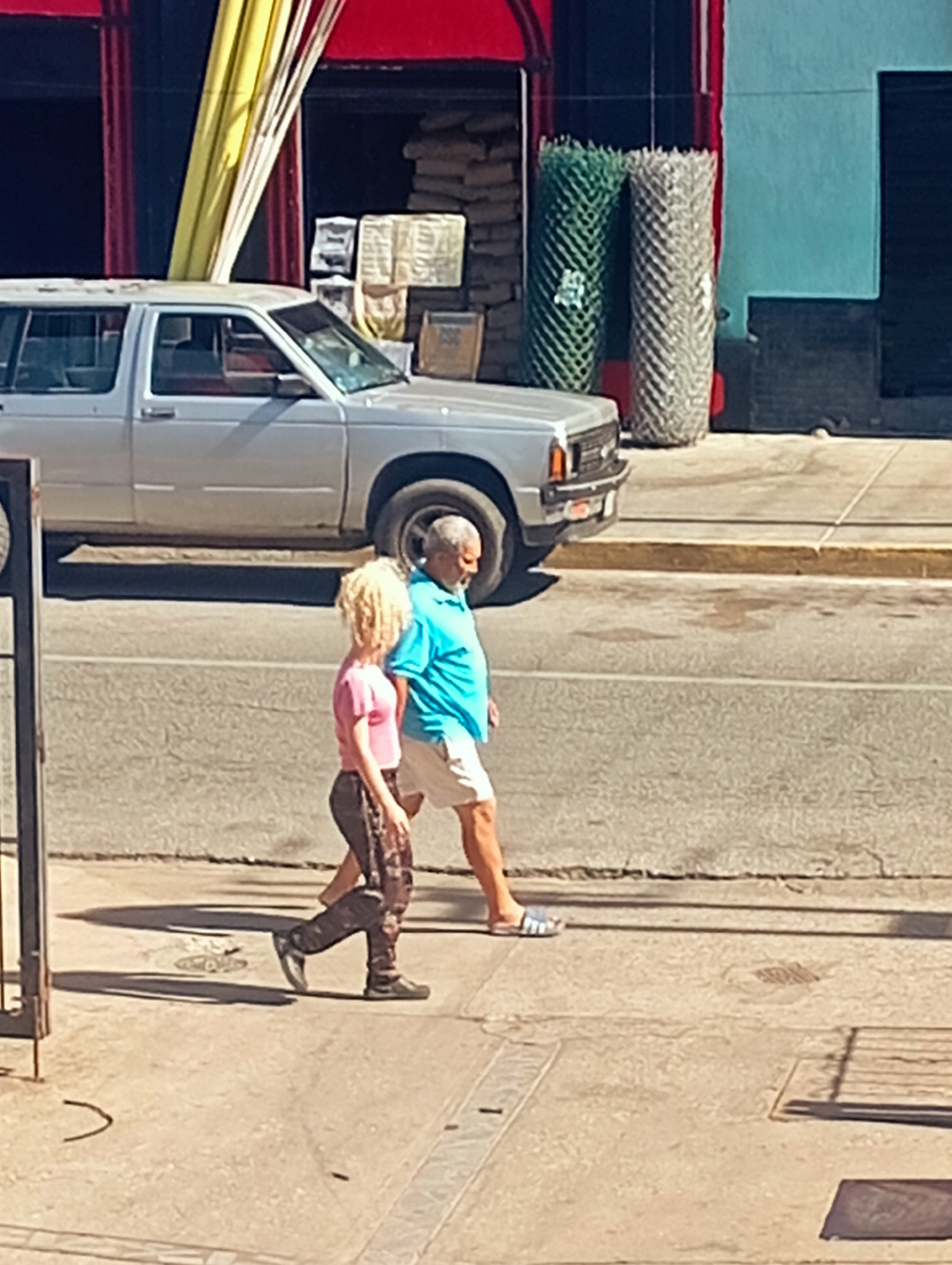 In Juan Griego, Venezuela, on a sunlit day, two individuals are walking on a cracked sidewalk next to a street. A man wearing a bright blue t-shirt and white shorts walks to the right, facing away from the camera. Beside him, a woman with voluminous blonde curly hair, wearing a pink t-shirt and patterned pants, also walks to the right, slightly ahead of the man and facing in the same direction. Behind them, a silver SUV is parked on the street. In the background, a red-and-black building facade is visible, with large yellow fabric draped across the top. Further back, large rolls of green and grey fencing material stand upright. The scene suggests a casual urban environment with elements of construction or storage visible.