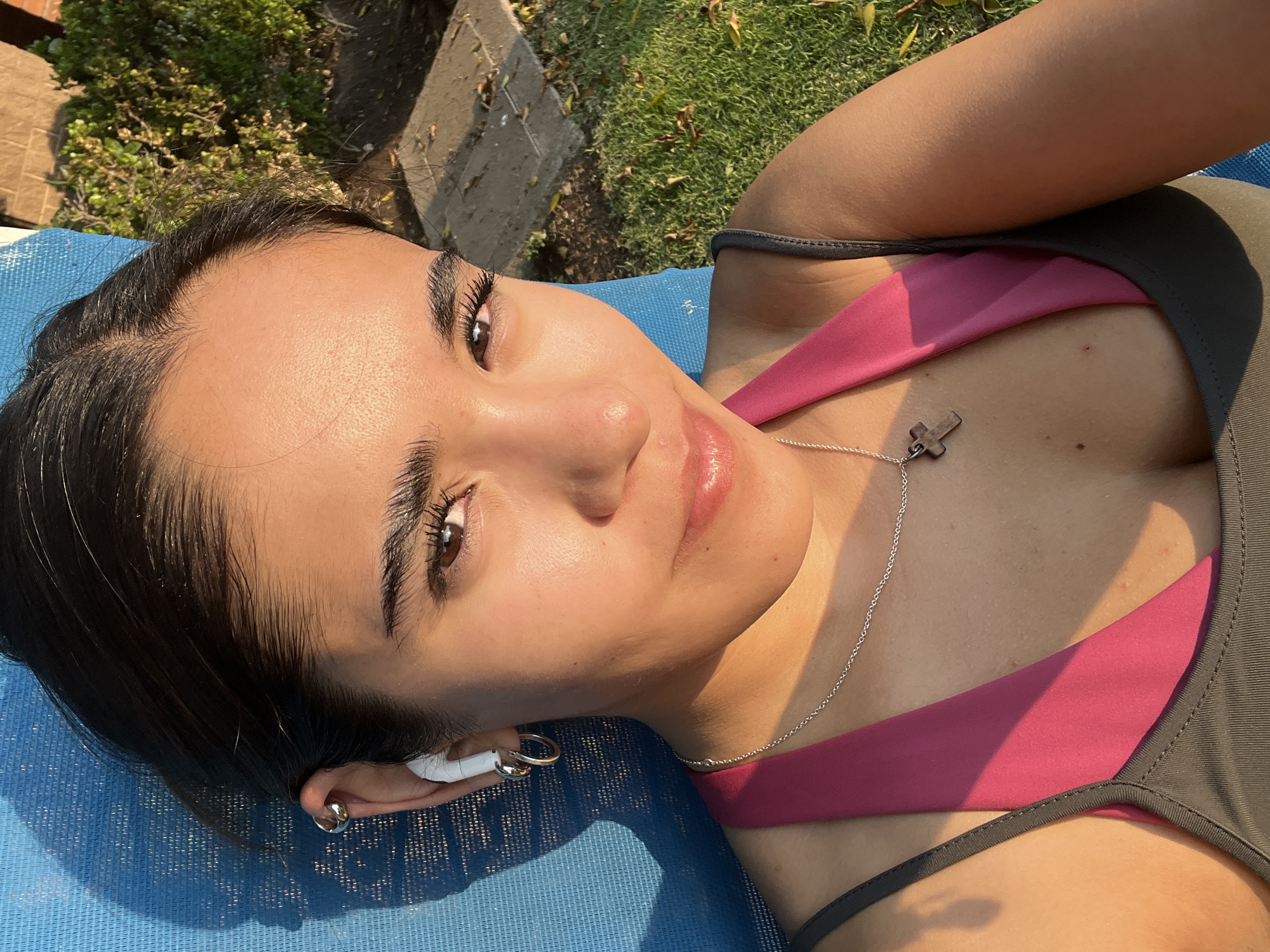 The image depicts a young woman lying on a blue surface, likely a mat or cushion, outdoors in what appears to be a sunny area. She is wearing a gray and pink workout top and a silver necklace with a cross pendant. A white earbud is visible in her left ear. Her hair is dark and pulled back. The background includes greenery, suggesting a garden or park setting. The specific location of San Bernardino Tlaxcalancingo, Mexico cannot be verified from the image alone.