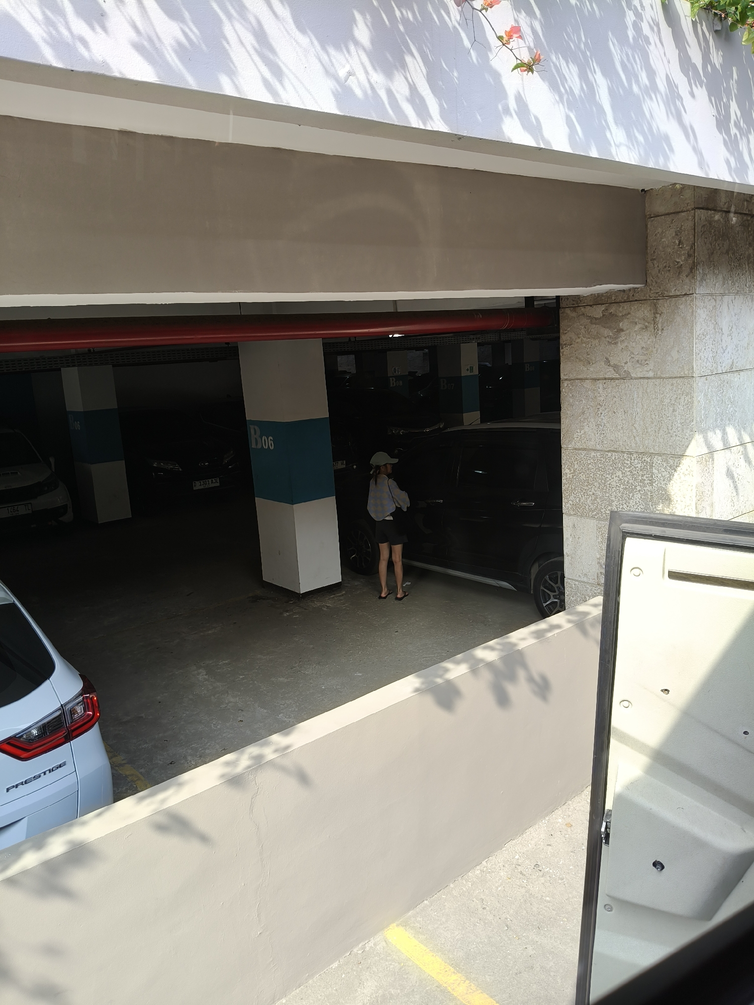 The image shows a parking garage with several cars. A woman in a hat and shorts stands next to a black SUV. The parking garage has concrete pillars with blue and white markings. Shadows from trees are cast on the ceiling. A white car with "PRESTIGE" written on the back is parked in the foreground. The scene appears to be in Desa Pagandaran, Indonesia, as indicated by the license plate of one of the cars, "D 3303 AJE", which is a common format for vehicle registration plates in Indonesia, specifically the "D" prefix often designates West Java, where Desa Pagandaran is located.