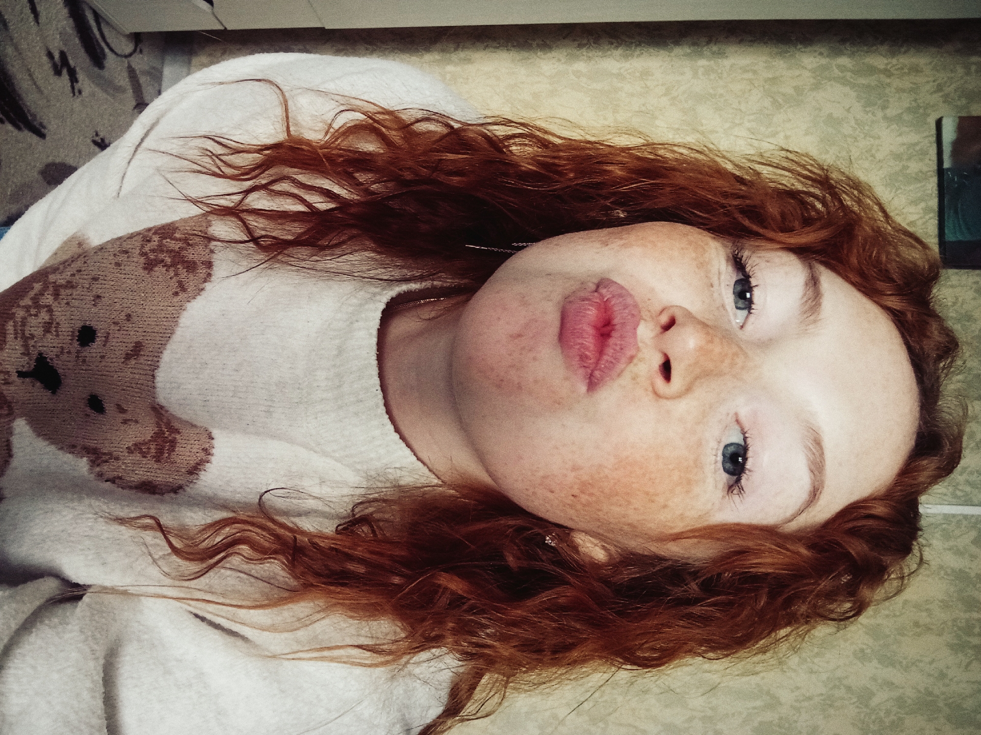 A close-up portrait captures a young person, likely female, with fair skin and numerous freckles across her nose and cheeks. She has vibrant red, wavy hair cascading around her, and striking light blue eyes. Her lips are playfully pursed in a "kiss" expression. She is wearing a light-colored (off-white or cream) knitted sweater adorned with a large brown teddy bear graphic on the left chest/shoulder area.
The subject is depicted lying down, with her head slightly tilted, looking directly at the camera. The setting is indoors; a dark-patterned rug or carpet is visible on the left, while a light-colored, patterned wallpaper covers the wall on the right. A small, dark rectangular object, possibly a framed item, is partially seen in the upper right background. The lighting suggests an indoor environment, and no specific time of day is discernible. The overall emotion conveyed is playful and casual. No text is visible in the image. The location context is Yurgamysh, Russia.