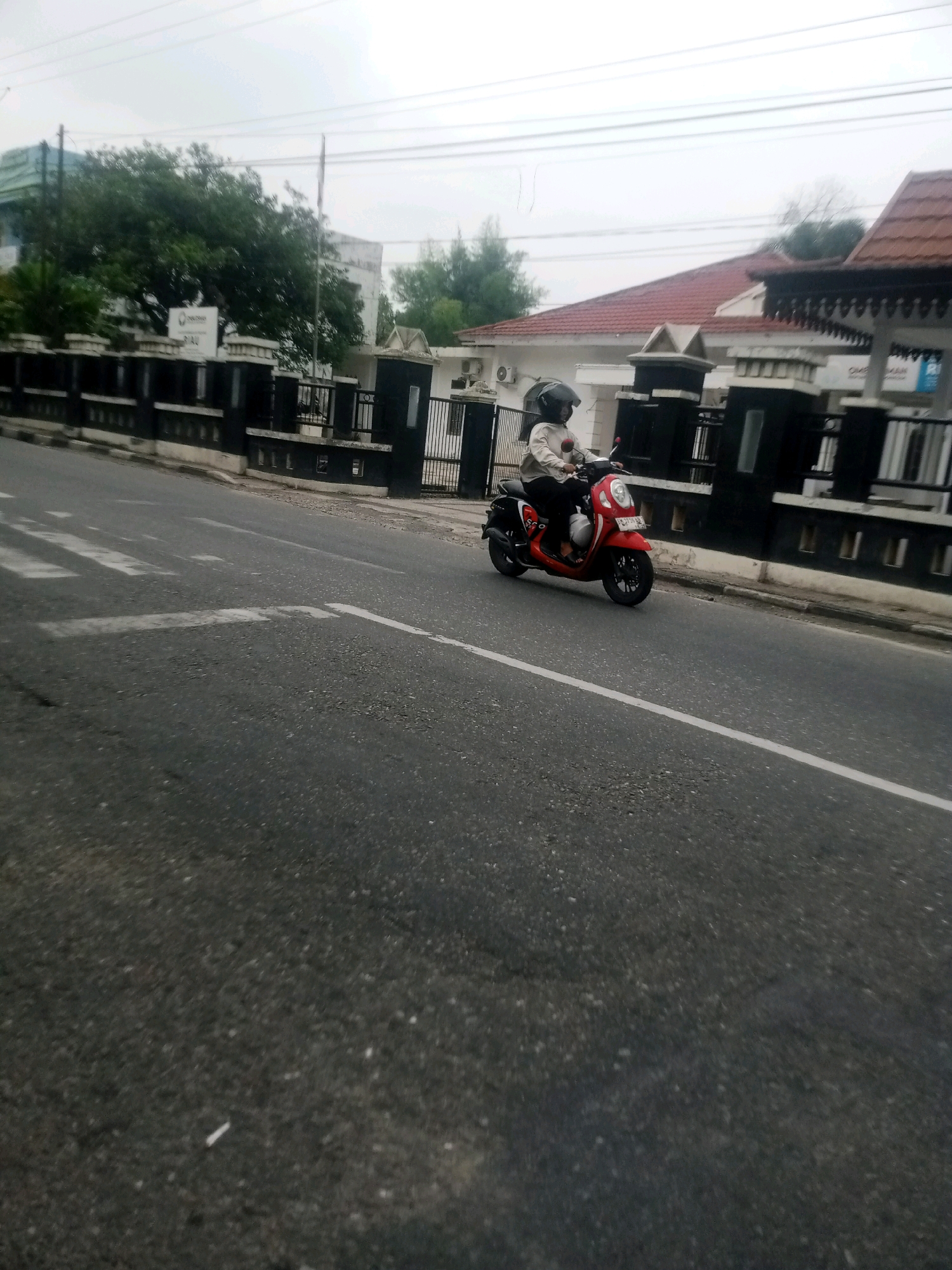 In Pekanbaru, Indonesia, a person wearing a helmet and a light-colored shirt rides a red scooter down a paved road. The scooter is moving towards the right side of the frame. The road has white lane markings. In the background, there is a black fence with ornate pillars. Behind the fence are buildings with red tile roofs and white walls, as well as trees and utility poles. The sky is overcast.