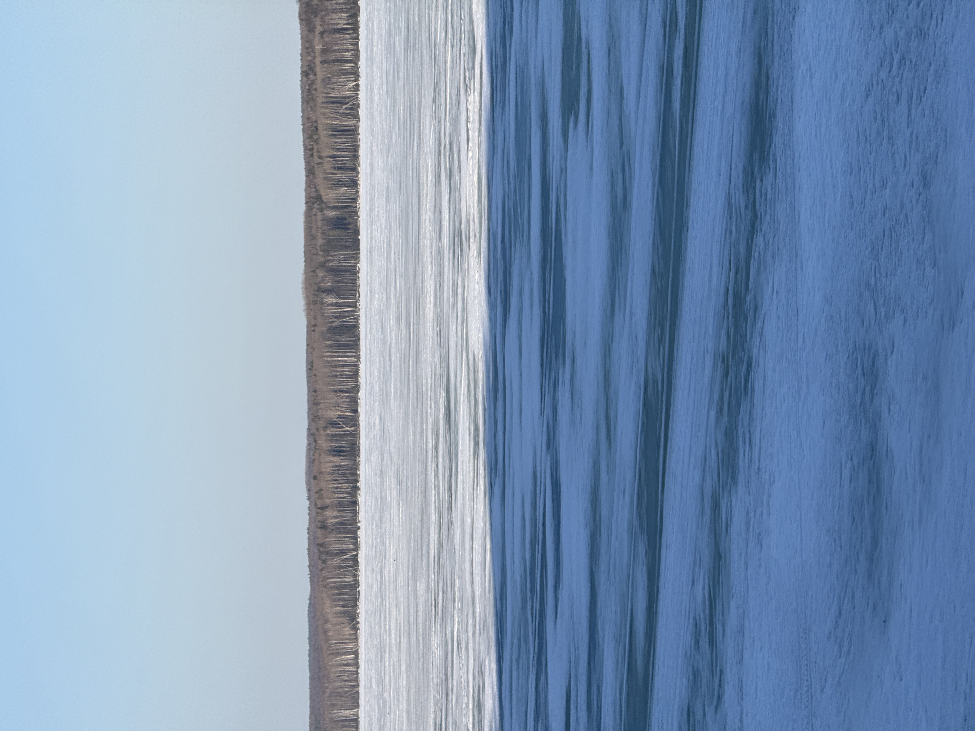 The image presents a view of the Kama River in Chaikovsky, Russia. From left to right, the image begins with a pale blue sky transitioning to a strip of land covered in dried vegetation, followed by the shimmering, light-reflecting surface of the Kama River, which darkens in tone as it extends to the right of the image. The texture on the river's surface indicates the presence of waves or ripples.