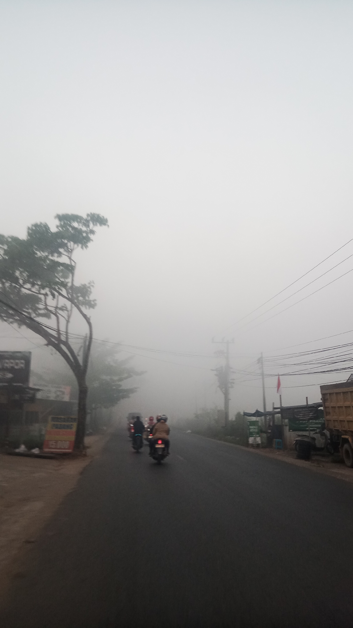 The image captures a street scene in Sungai Sipai, Indonesia, shrouded in heavy fog. A paved road, wet with mist, is visible, with motorcycles and their riders traveling along it. The fog obscures much of the background, creating a hazy atmosphere with limited visibility. On the left side of the road, a building with signs, one with the text "RAPPOR", is partially visible, along with a tree. Further down the road on the right, there are several structures, including what appears to be a truck and other vehicles. A red and white flag is displayed on a pole. The overall impression is one of a damp, overcast morning in a rural or suburban area.