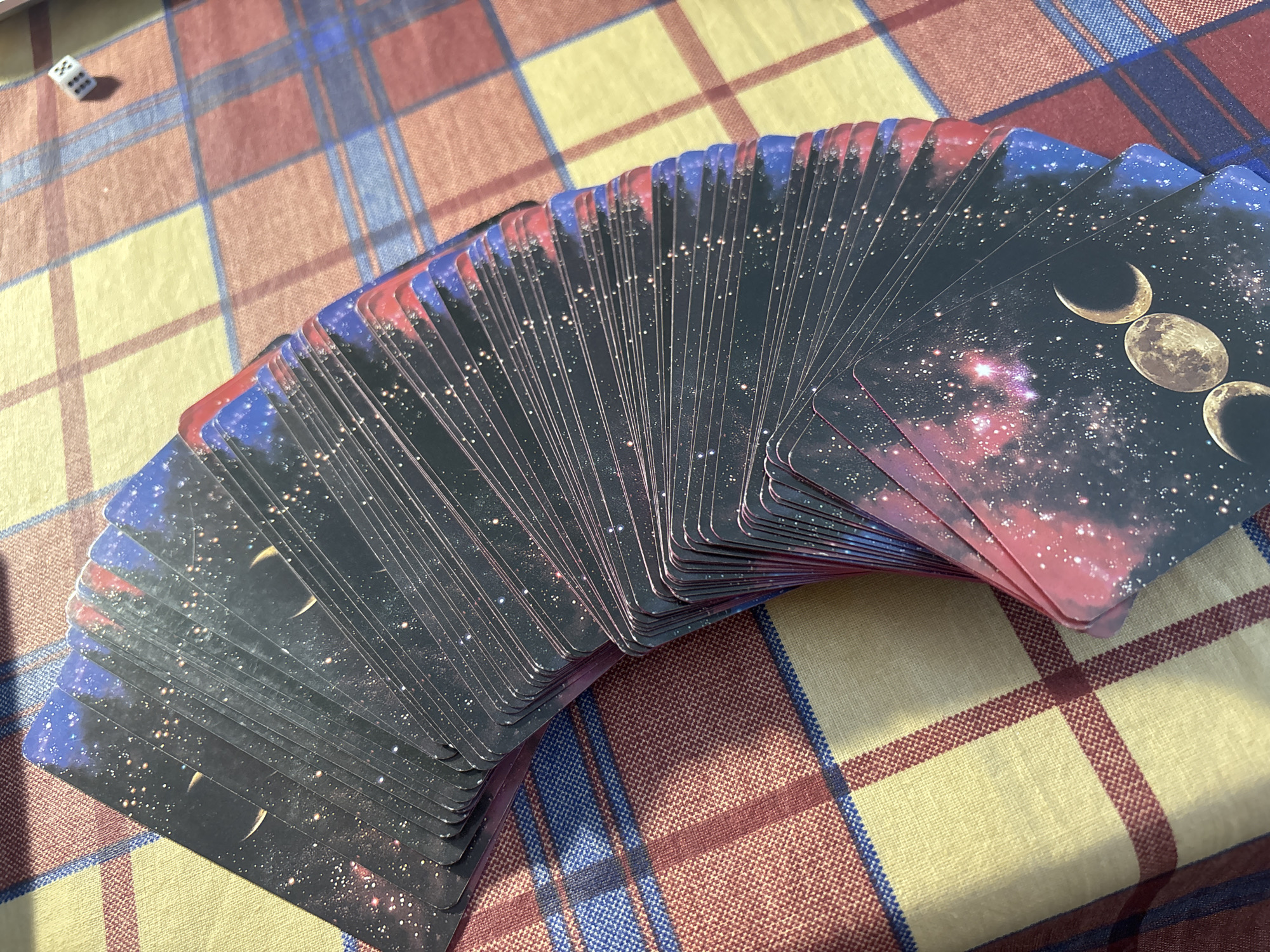 A spread of tarot cards with a celestial design is fanned out on a checkered tablecloth. The cards feature a dark background with numerous stars and a depiction of moon phases. The tablecloth has a pattern of yellow, red, and blue stripes. In the upper left corner of the image, a single six-sided die rests on the tablecloth. The lighting suggests it is daytime, and the setting appears to be indoors. No people are visible in the image, and no text is present on the cards or the tablecloth.