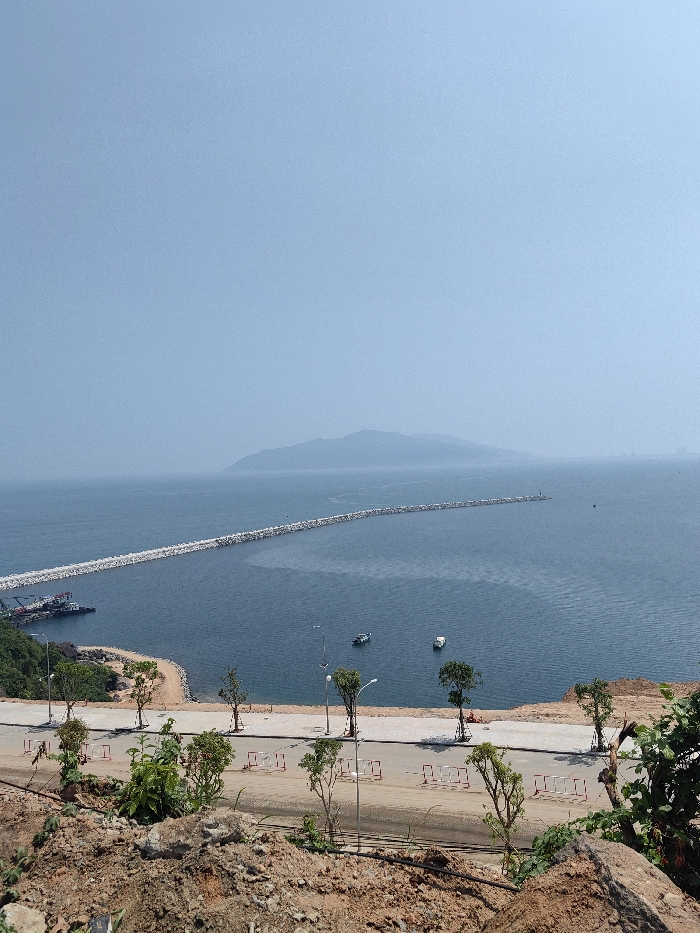 An elevated, wide-angle view captures a coastal scene in Hải Vân Ward, Vietnam. The foreground is composed of reddish-brown earth, rocks, and sparse green vegetation. A newly constructed paved road, bordered by white and red barriers and lined with young trees and streetlights, traverses the scene horizontally below the viewpoint. This road meets a small, light-colored sandy beach and a cove on the left, where a small port facility with two moored industrial vessels is visible. Extending into the calm, dark blue bay water is a long, straight breakwater, constructed from numerous grey rock formations, which stretches from left to right. Two small, light-colored boats are positioned on the open water. In the distant background, a hazy mountain range or island is visible on the horizon, merging with the pale blue sky. Faint, indistinct structures are discernible on the distant coastline to the far right. No individuals are visible.