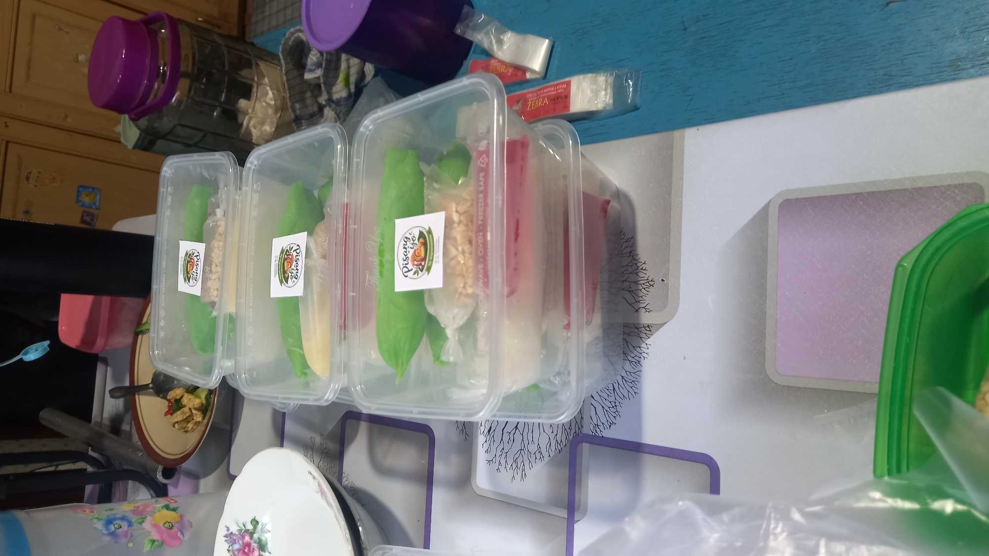 The image depicts a collection of takeaway food containers, likely prepared for delivery or personal consumption, set on a table. The containers are transparent plastic, stacked vertically. The contents of the containers appear to be a sweet treat, possibly some type of pastry or dessert, wrapped in green-colored material and some contain a creamy white substance and possibly peanuts. A sticker with the text "Pisang Yo" is visible on several of the containers, indicating the brand or name of the food. Other items on the table include a ceramic bowl with food, a water pitcher, and a purple-lidded container. The overall setting suggests a domestic environment, possibly a kitchen or dining area. The presence of a blue wooden table and a patterned tablecloth adds to the informal atmosphere. While the exact location is not specified, such a scene is common in residential areas of cities like Balikpapan, Indonesia, where home-based food businesses are prevalent.