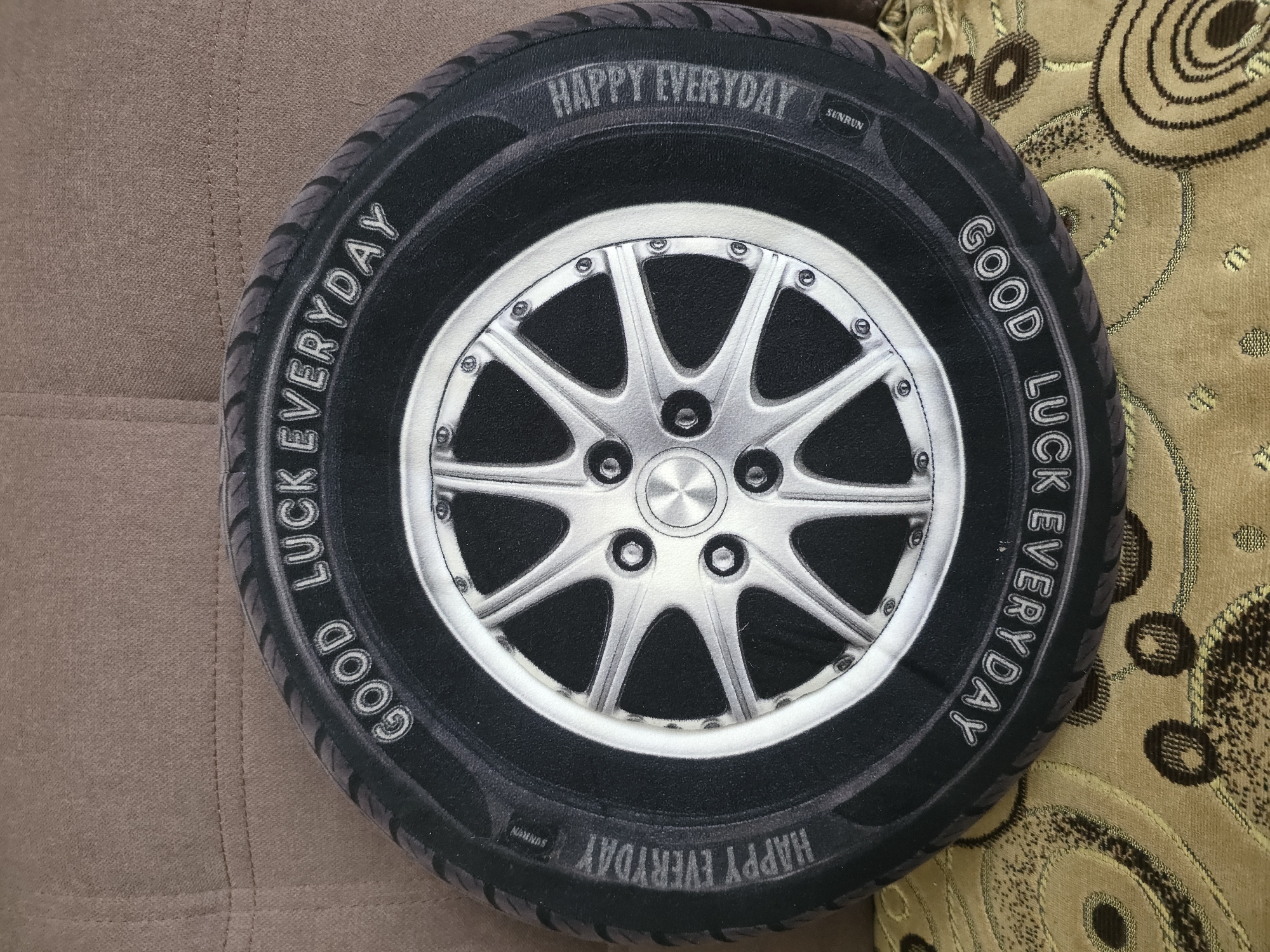 The image shows a round pillow designed to look like a car tire with a silver rim. The tire has white lettering around its circumference. On the top portion of the tire, the words "GOOD LUCK EVERYDAY" are visible. On the bottom portion of the tire, the words "GOOD LUCK EVERYDAY" are also visible, but appear upside down from this perspective. The pillow rests on a textured, brown upholstered surface. The pillow itself is detailed, with a realistic depiction of tire tread and a shiny rim with lug nuts. The background is a plain brown fabric, possibly a couch or chair. No people are present in the image. The specific location in Qarabalıq, Kazakhstan is not discernible from the visual information provided.