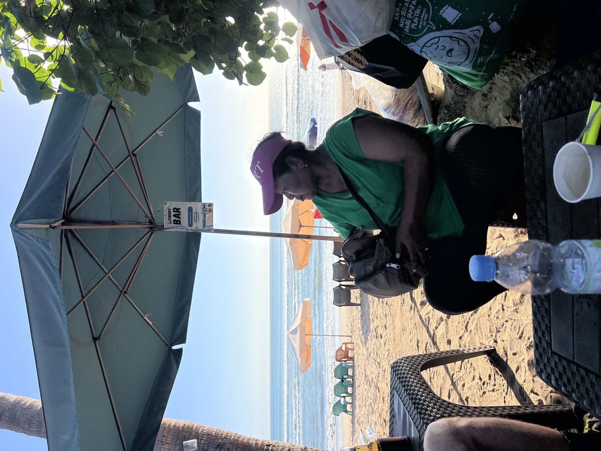 A woman in a green t-shirt and a pink visor sits at a beachside cafe in Kuta, Indonesia, under a large umbrella. She is looking out at the ocean, holding a dark bag. In front of her are tables and chairs on the sandy beach, with the ocean waves and several beach umbrellas in the background. A bottle of water and a cup are on a table. The scene appears to be a relaxed day at the beach.
