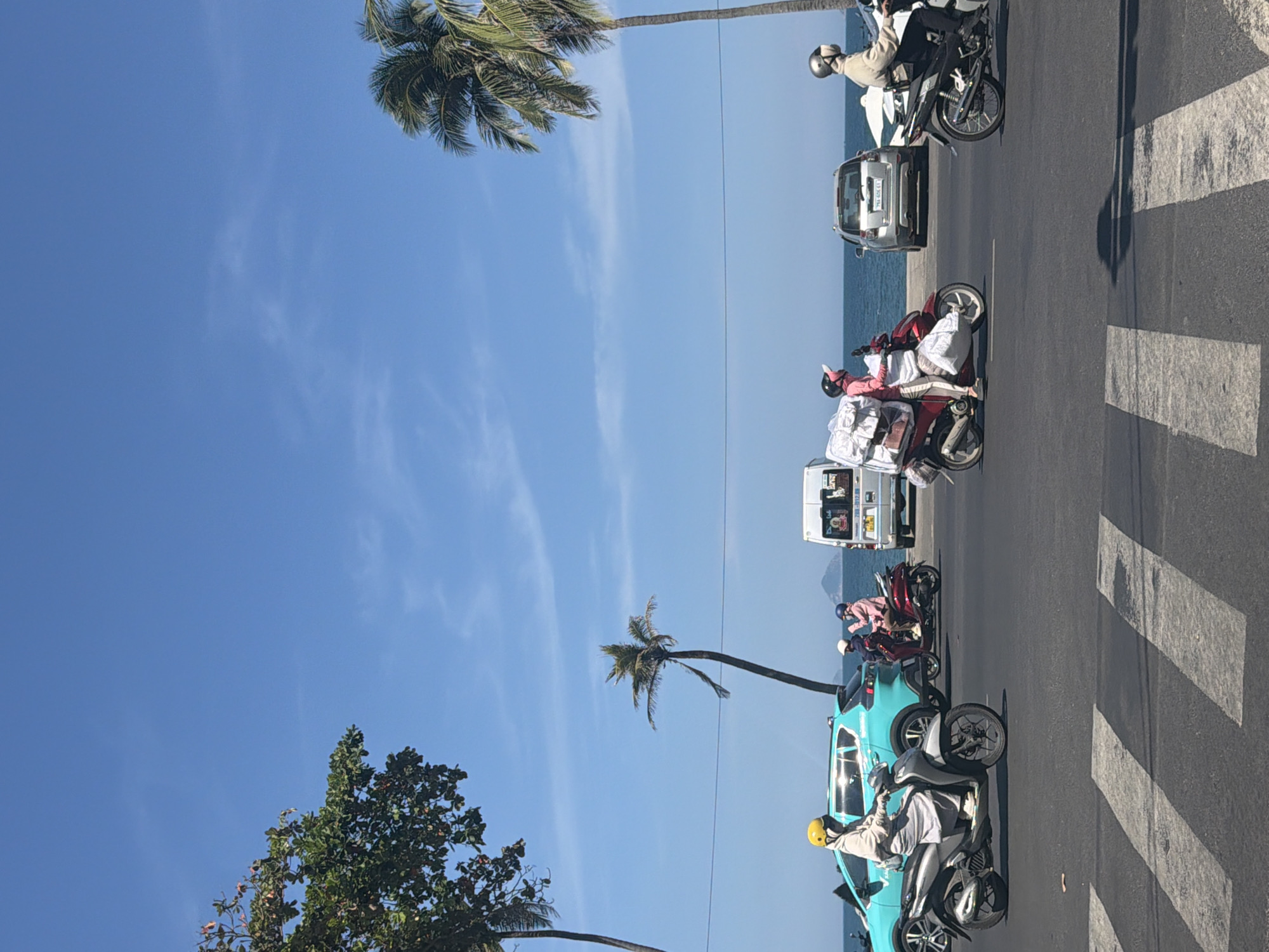 This image depicts a street scene in Nha Trang Ward, Vietnam, characterized by a bright, clear blue sky and the presence of palm trees and vegetation. The foreground is dominated by a paved road with a white pedestrian crossing. Several people on motorcycles are traveling along the road.
On the left, a person wearing a yellow helmet and a light-colored outfit rides a silver scooter, with a turquoise SUV to their left. Further down the road, two individuals on a red scooter, one with a pink helmet and the other with a white helmet, are carrying a large load of white plastic-wrapped items.
In the background, a white van is visible, and beyond that, the sea stretches out under the blue sky. Two cars are parked on the side of the road near the sea, and another person on a black motorcycle is seen moving away from the camera on the right side of the road. The overall atmosphere is that of a sunny day in a coastal urban environment.