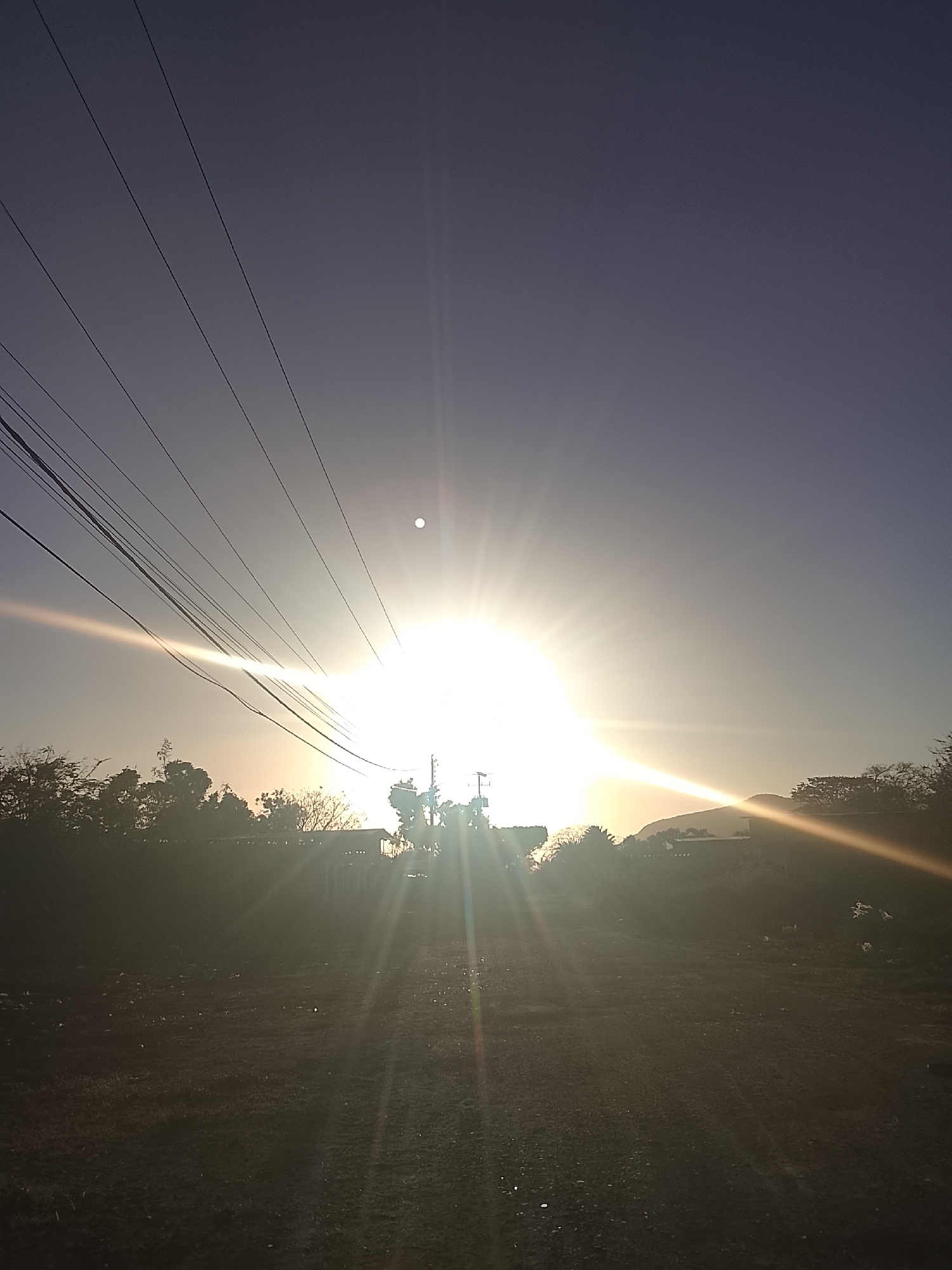 The image depicts a brightly illuminated scene at or near dusk or dawn. The light source, likely the sun, is intensely bright, positioned centrally in the upper portion of the frame, causing significant lens flare and radiating light beams. A series of power lines stretch across the upper left quadrant of the image. Below the light source, the foreground is predominantly dark, revealing a field with low vegetation. Silhouetted against the bright light are trees and what appears to be buildings. In the background, a hill or small mountain is discernible on the right side of the image, partially obscured by the glare. The sky transitions from a lighter hue near the horizon to a darker blue towards the upper regions of the frame. The image has no visible people or animals. The location, based on the prompt, is Altagracia, Venezuela.