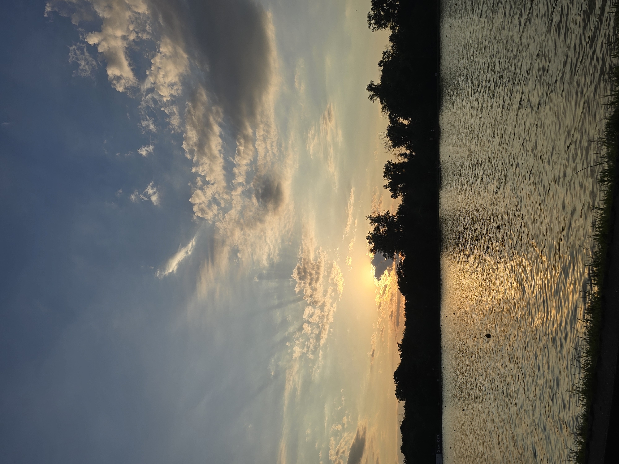 A sunset over a tranquil body of water with a silhouetted tree line on the left. The sky is a canvas of soft blues and oranges, with wispy clouds catching the fading sunlight. Rays of light emanate from behind the clouds, creating a dramatic effect. The water reflects the warm hues of the sunset, with ripples distorting the golden light. The dark silhouette of trees lines the left edge of the frame, suggesting a natural landscape. The overall mood is peaceful and serene, evoking a sense of calm as the day draws to a close. The image appears to be taken in Plovdiv, Bulgaria. The time of day is sunset.