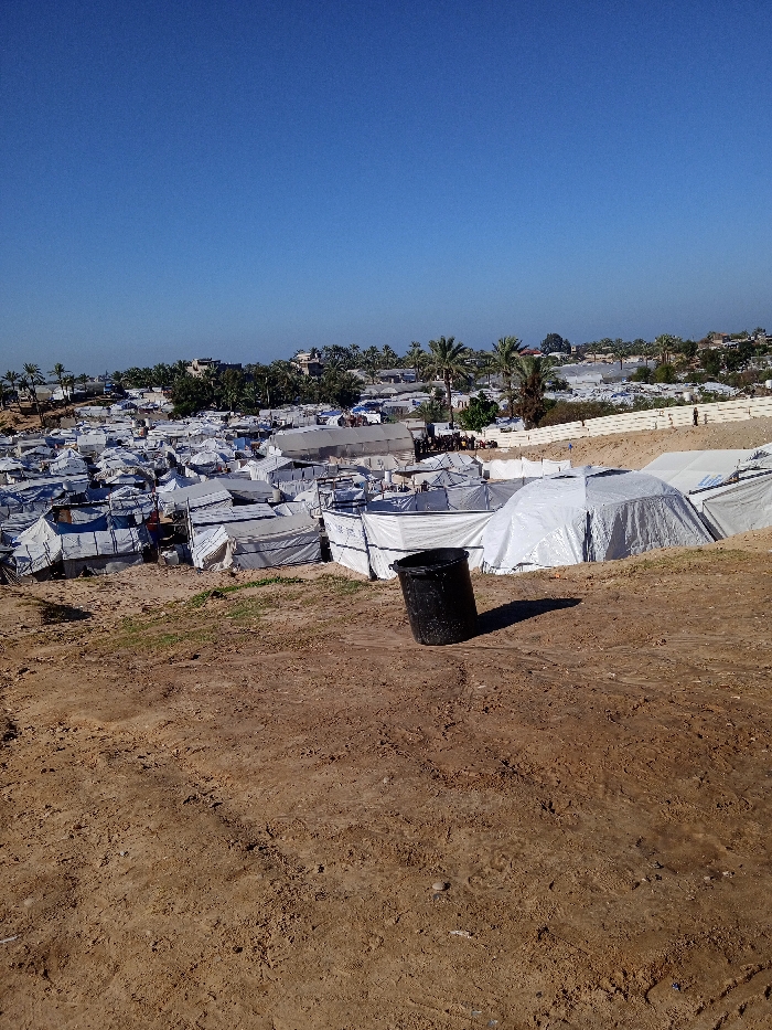 An outdoor, wide-angle shot captures a densely packed temporary shelter encampment under a clear, cloudless blue sky, identified as being in Khan Yunis, Palestinian Territory. The foreground consists of dry, reddish-brown earth with minimal vegetation. A single black plastic bucket stands on the bare ground, casting a short shadow to its right, indicating a high sun angle. The midground is dominated by a sprawling arrangement of numerous makeshift shelters and tents, predominantly constructed from white or light-colored tarpaulins, tightly clustered together. Some tents exhibit a dome shape, while others are more rectangular, with at least one structure displaying a visible UNRWA logo. In the background, a line of palm trees and other foliage delineates the extent of the camp, with additional buildings visible amongst the trees. A light-colored barrier or wall is present on the right side of the background, with a few small, indistinct human figures visible near it in the distance. No specific actions or interactions are discernible among people within the visible parts of the camp.