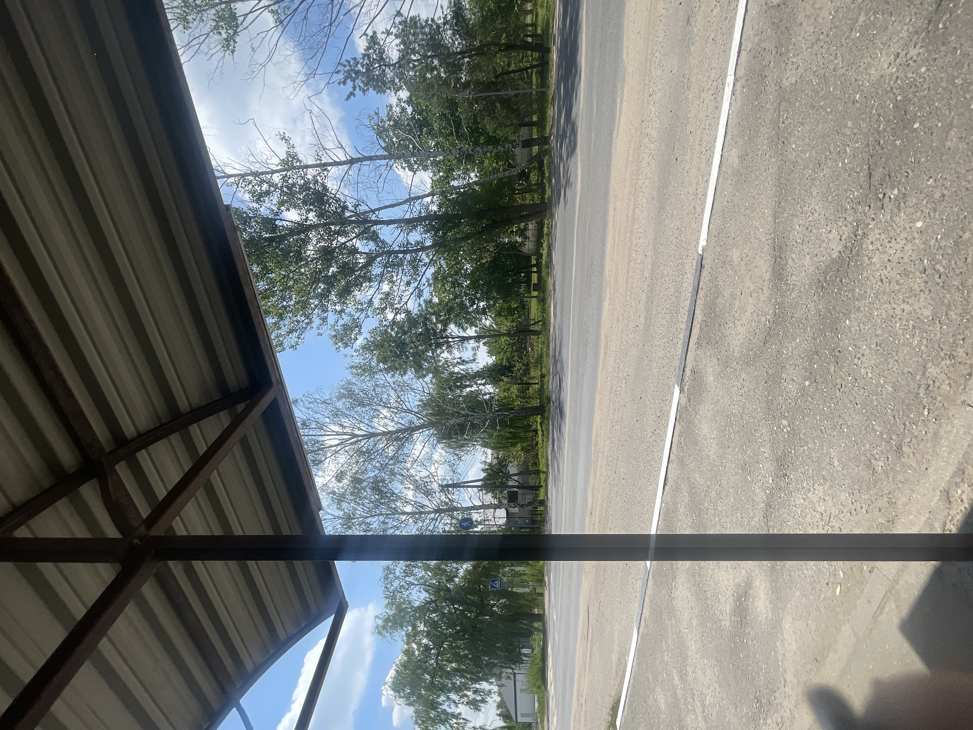 The image shows a view from under a corrugated metal roof supported by a dark metal frame, looking out onto a paved road. A single, dark vertical support post is visible in the foreground, casting a shadow. The road is mostly gray asphalt with some texture and white markings indicating a lane divider or pedestrian crossing. Beyond the road, a line of trees with green foliage borders the scene. In the distance, there are some buildings and more trees. The sky is blue with scattered white clouds. A pedestrian crossing sign and a blue circular sign with a white pictogram are visible among the trees on the left side of the road. The overall environment appears to be a rural or semi-rural setting, possibly along a road in the vicinity of Мухавецкі сельскі Савет, Belarus.