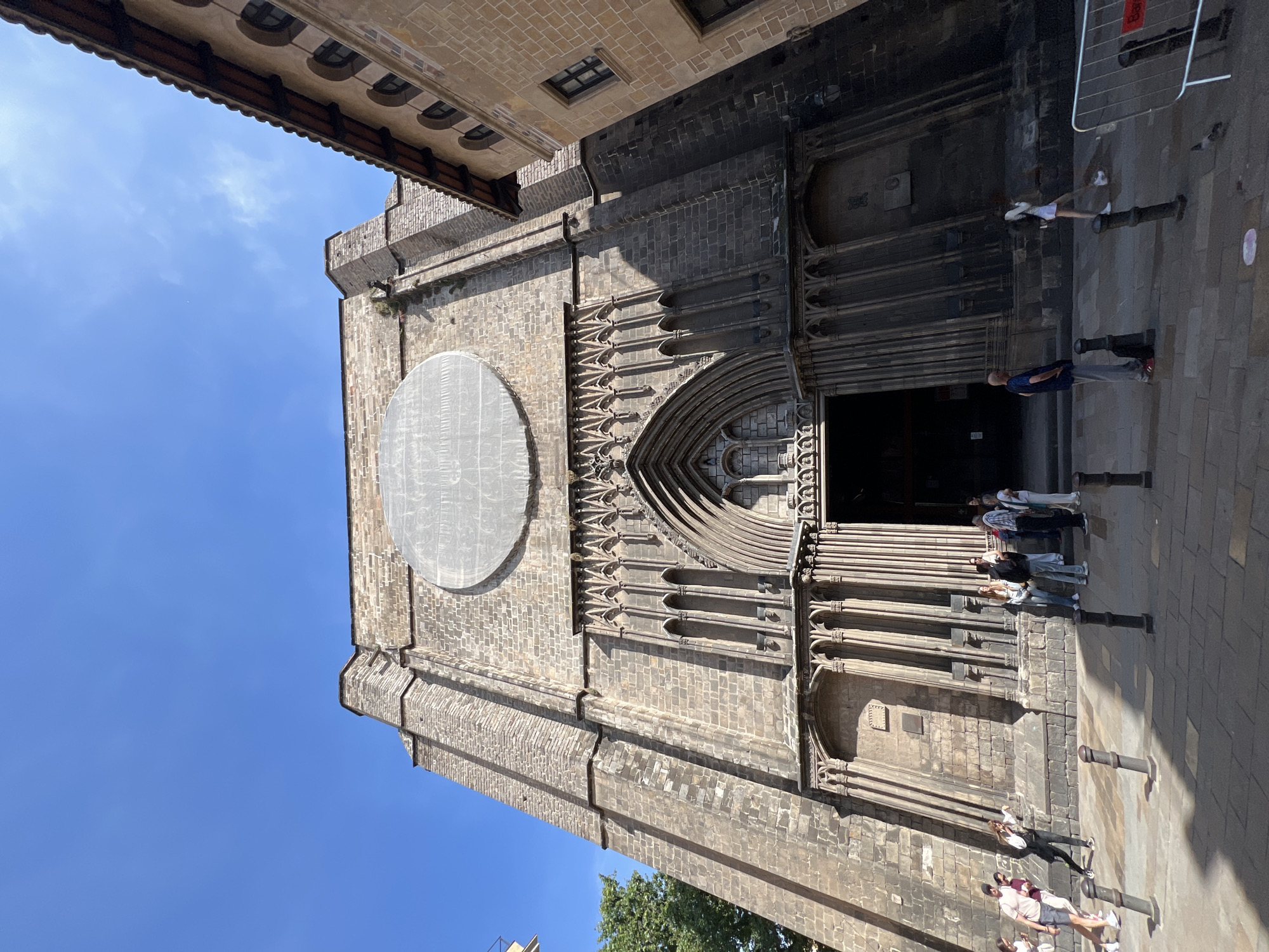 The image captures the facade of the Basilica of Santa Maria del Pi in Barcelona, Spain, a Gothic-style church constructed in the 14th century. The lower part of the facade shows the church entrance, along with several people standing in front of it. The building's structure is made of dark stone, with a large circular feature at the top. The sky is bright blue. A small portion of another building can be seen above the church, with windows and decorative elements.