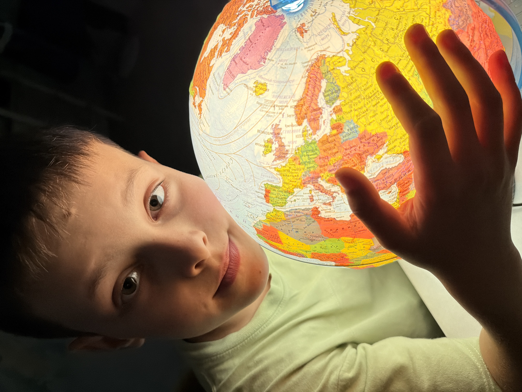 A young boy, appearing to be around 8-10 years old, is shown in a close-up shot, holding a brightly lit globe. He is wearing a pale green t-shirt. His face is partially illuminated by the globe, casting a warm glow on his skin. His eyes are looking towards the viewer, with a curious and thoughtful expression. The globe itself displays colorful continents and bodies of water, with text labels in Cyrillic script, indicating it's likely a Russian-language globe. His right hand is gently placed on the globe, with his fingers partially obscuring some of the landmasses. The background is dark and out of focus, creating a sense of intimacy and focus on the boy and the globe. The scene appears to be set indoors, possibly in a bedroom or study, given the lighting and the presence of a table or desk beneath the globe. There are no other people or objects clearly identifiable in the background. The image captures a moment of engagement and exploration with the world represented by the globe.