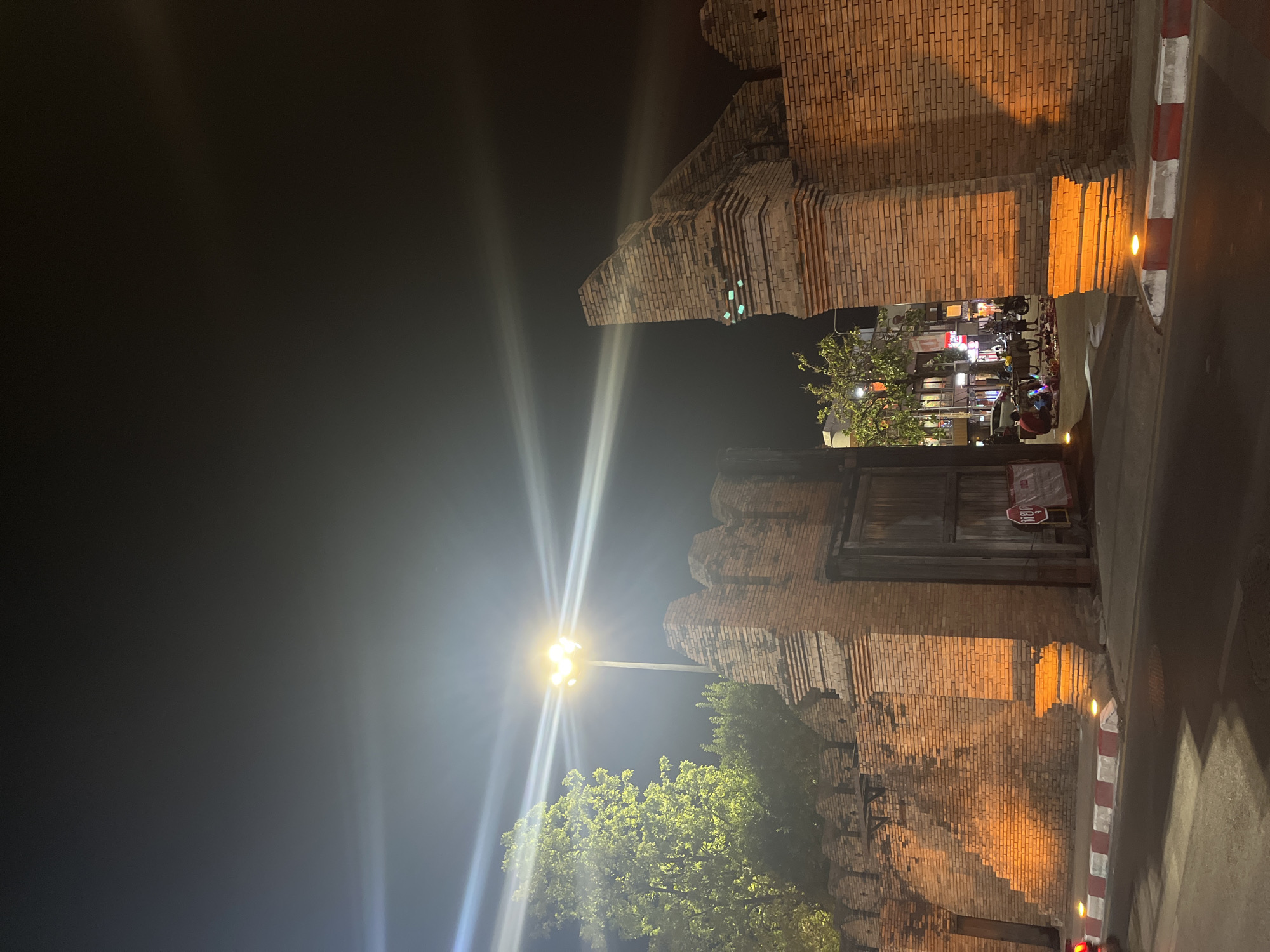 The image shows the Tha Phae Gate in Chiang Mai City Municipality, Thailand, at night. The brick structure of the gate is visible, illuminated by streetlights. A street runs alongside the gate, with a red and white curb. In the background, lights suggest activity in the surrounding area. The sky is dark.