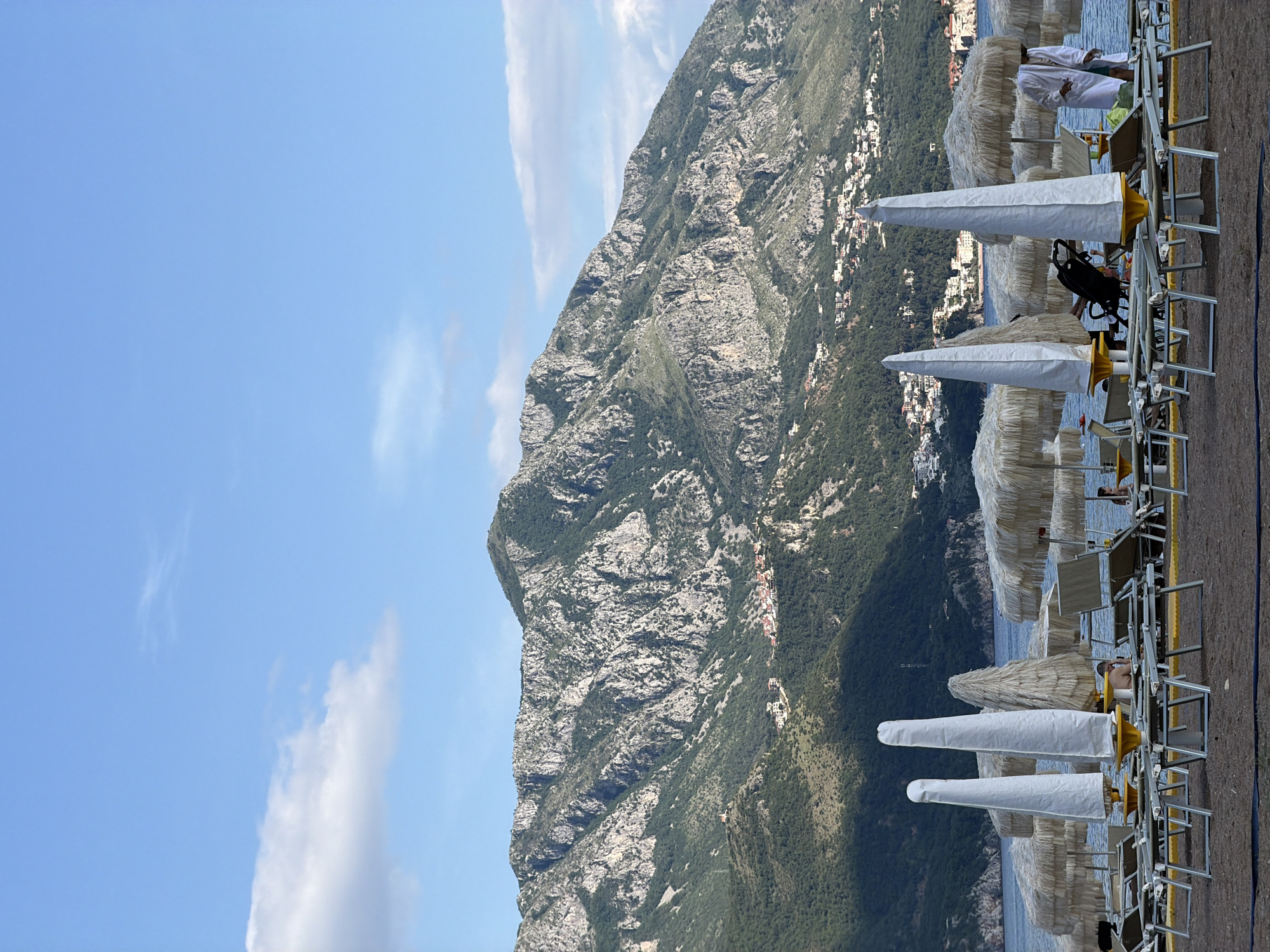 The image depicts a coastal scene, likely a beach resort in Boreti, Montenegro, juxtaposed with a steep, rocky mountain. In the foreground, a row of beach chairs and white sun umbrellas lines the edge of a sandy area, presumably a beach. People can be seen reclining in some of the chairs, and a few are visible walking or standing. The water extends beyond the beach, leading to the base of the mountain. The mountain occupies a large portion of the image and appears rugged with sparse vegetation and rocky outcroppings. Dotted along the mountain's lower slopes are structures that suggest a town or village. The sky above is blue with some scattered clouds.