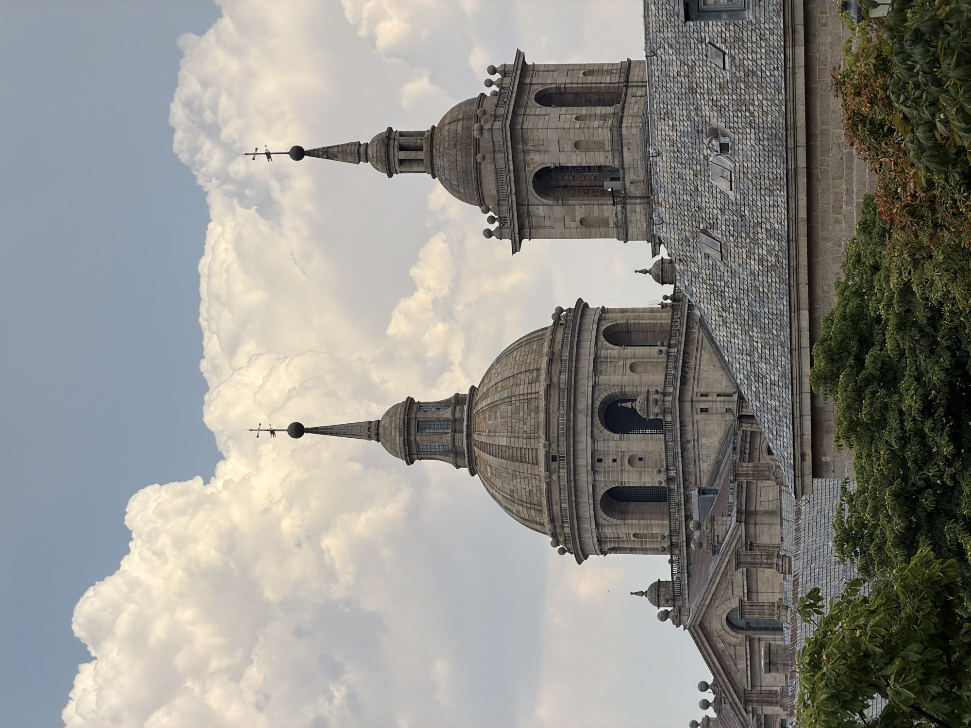 This media file captures a portion of the Monastery of San Lorenzo de El Escorial in Spain. The primary subjects are the impressive stone domes and spires of the monastery, characteristic of Renaissance architecture. The scene is an outdoor view, focusing on the upper sections of the buildings.
The image appears to be taken during the daytime, with soft, diffused lighting suggesting it could be morning or late afternoon. The weather seems fair, with a mix of clear blue sky and large, fluffy white clouds dominating the background on the left. The right side of the frame shows lush green trees and foliage, hinting at a warm climate and possibly a temperate season.
Notable details include the intricate stonework of the monastery, the tiled roof, and the weather vane perched atop one of the spires. The presence of the natural landscape surrounding the historical building provides a sense of scale and context. There is no visible text in the image. The overall mood is one of grandeur and historical significance, with the natural elements offering a sense of tranquility.