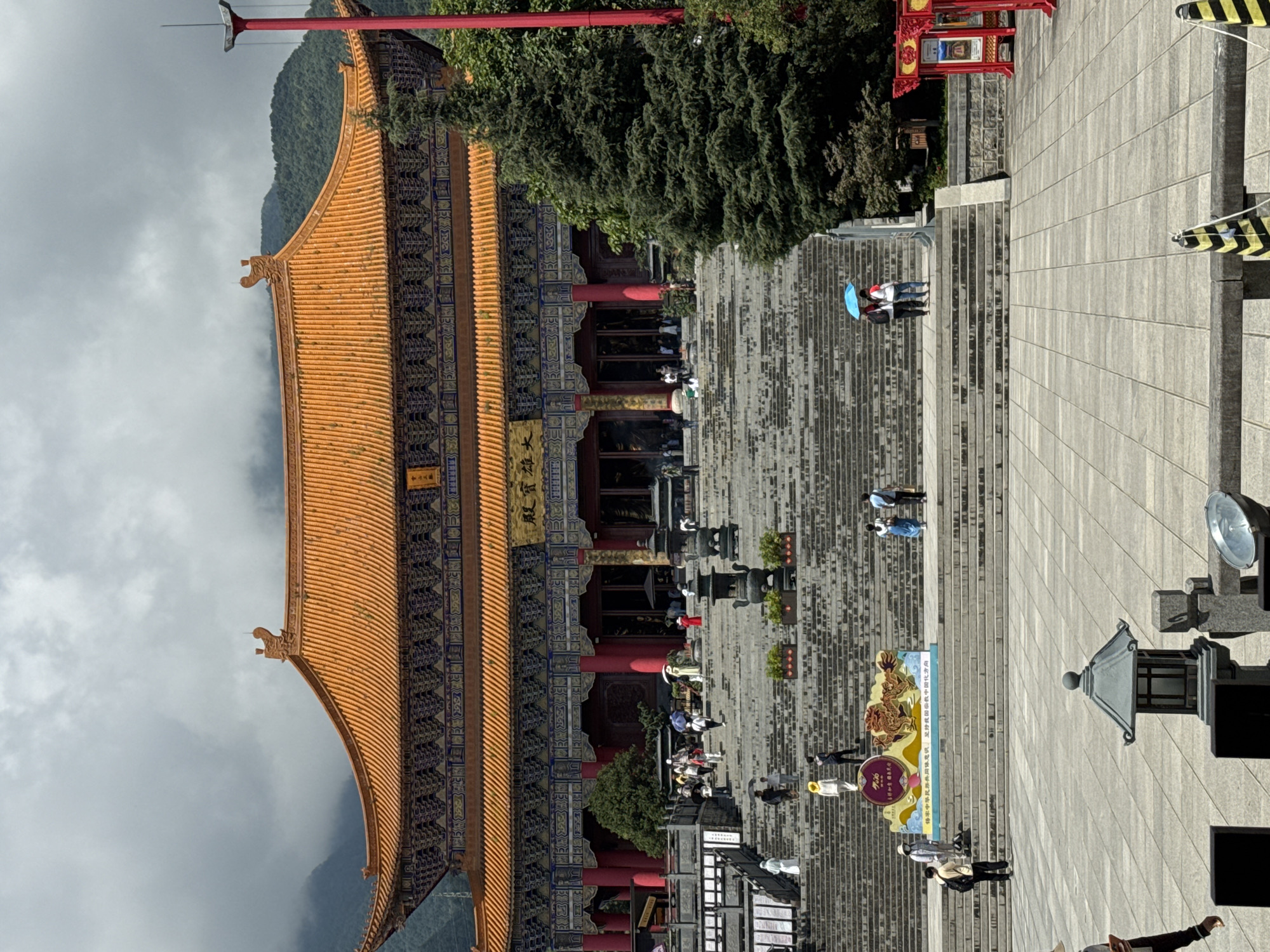 The image depicts the entrance to the Three Pagodas of Chongsheng Temple in Dali City, China. A grand temple structure with an orange tiled roof and intricate blue and gold detailing dominates the upper half of the frame. A wide, imposing stone staircase leads up to the temple entrance, with numerous people ascending and descending. Several large incense burners are placed on the steps. On the left side of the staircase, a colorful banner featuring a golden dragon is displayed. The lower part of the image shows a paved plaza with several stone lanterns and some decorative barriers. Green trees, including a tall conifer on the right, flank the staircase and temple. The sky is overcast with clouds, and a misty mountain range is visible in the background.