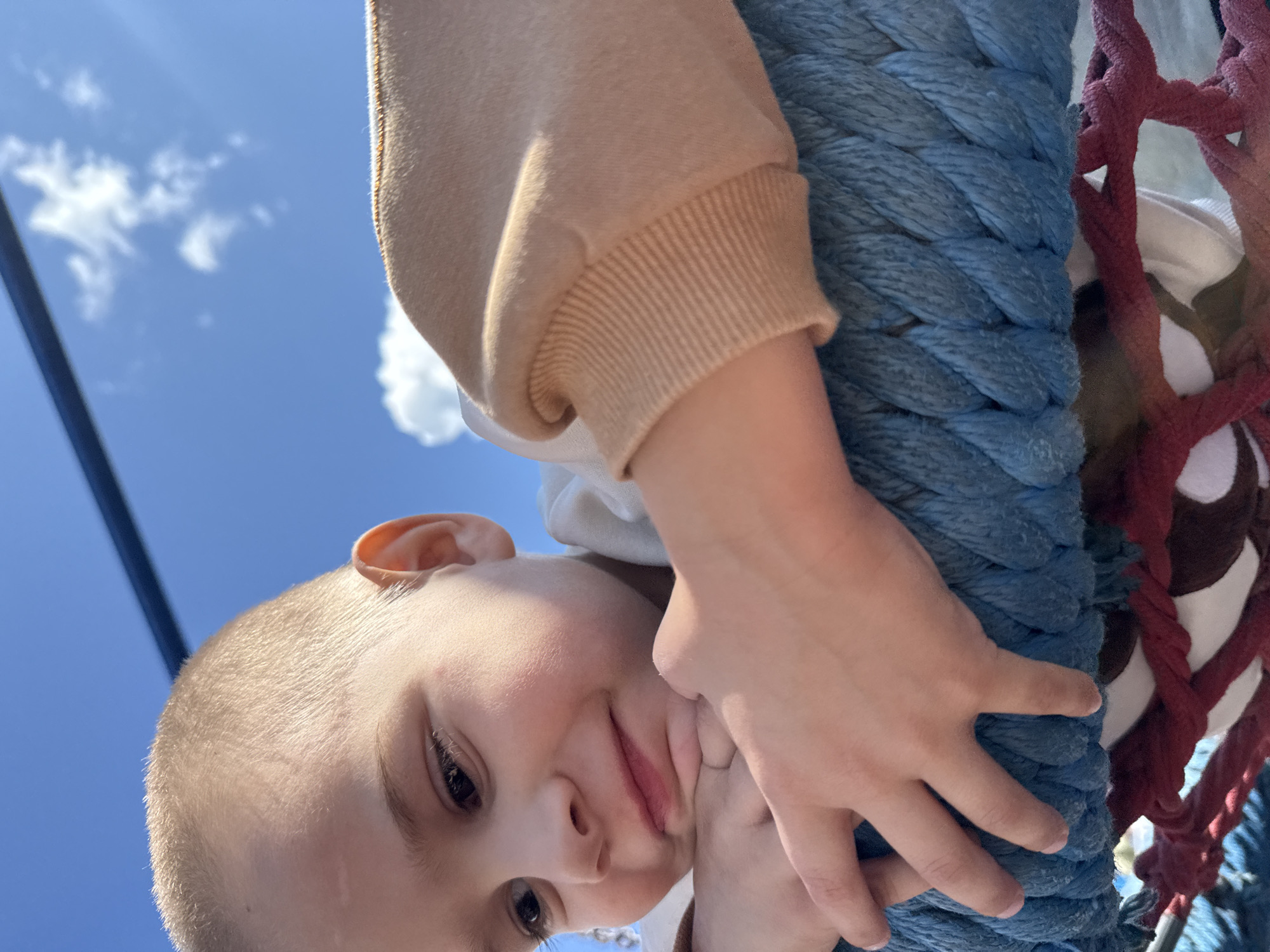 The image captures a close-up, low-angle shot of a young child resting their chin on their hands. The child has short, light-colored hair and appears to be looking down and slightly to the left with a gentle smile. They are wearing a light-colored shirt and a tan sweatshirt with ribbed cuffs. The child's left arm and hand are visible, resting on a thick, braided blue rope. Behind the child, a bright blue sky with scattered white clouds and a dark, thin horizontal line, possibly a railing or structure, are visible. The rope appears to be part of a playground or an outdoor recreational structure. There is no discernible information in the image to suggest a specific location within Gornozavodsk, Russia.