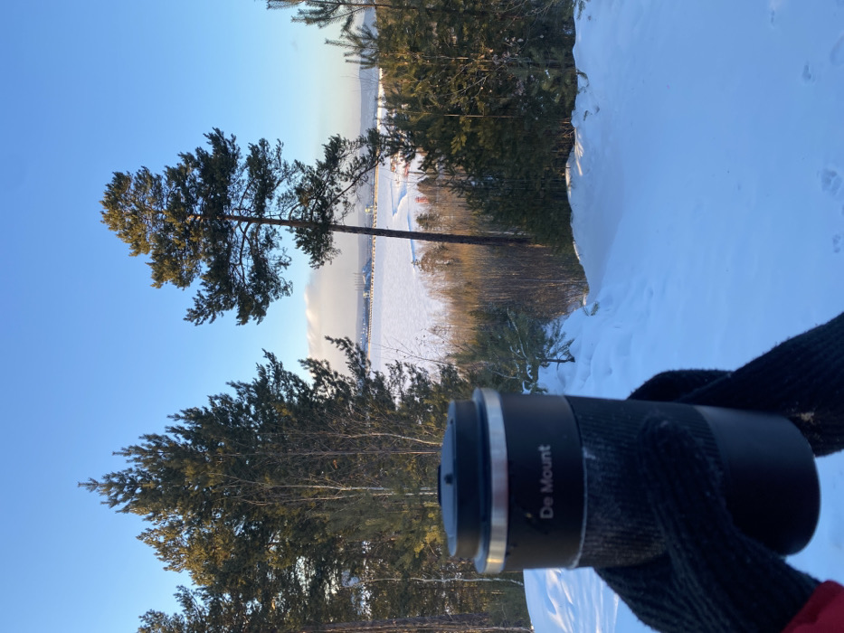 A forensic description of the scene depicts an outdoor winter landscape in Ust-Ilimsk, Russia, under a clear, bright blue sky. In the foreground, a person's left hand, clad in a black knitted glove with a subtle red cuff detail, holds a black thermal mug. The mug features a silver-colored rim, a black lid, and "De Mount" inscribed in white text on its upper body above a textured black grip. The immediate ground is covered in deep, undisturbed white snow, sloping downwards.
The midground features a dense forest composed primarily of tall pine trees with dark green needles and several bare deciduous trees. Sunlight illuminates parts of the forest, casting shadows. Beyond the forest, a wide expanse of frozen, snow-covered water is visible, consistent with the Ust-Ilimsk Reservoir or the Angara River.
In the background, on the far bank of the frozen water, a large industrial complex is present. This complex includes multiple substantial structures, possibly towers or cranes, and a long, linear structure extending across the water, indicative of a dam or a bridge. This configuration is consistent with the Ust-Ilimsk Hydroelectric Power Plant. The horizon above the industrial complex shows faint, wispy white clouds. The overall lighting suggests a cold, sunny day with a low sun angle. No other individuals or explicit actions are observed.