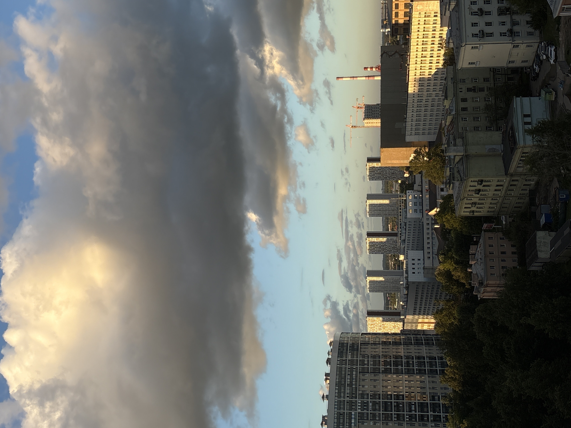The image captures a city skyline during what appears to be late afternoon or early evening, with dramatic cloud formations dominating the upper portion. The sky transitions from a bright, sunlit yellow and white at the top, indicative of sunlight filtering through clouds, to darker, grayer clouds lower down, with patches of blue sky visible.
Below the sky, a cluster of modern residential buildings stands out. The most prominent is a large, curved, light-colored building on the left, featuring numerous windows and a distinctive rooftop design with several raised structures. To its right, a row of taller, rectangular buildings with patterned facades is visible, some of which are illuminated by the setting sun, casting a warm glow. Further in the distance, more buildings of varying heights and architectural styles form the urban landscape.
On the right side of the image, industrial elements are present, including a tall, striped smokestack and construction cranes against the backdrop of buildings. The foreground is filled with dense, dark green trees, obscuring the lower sections of some buildings and creating a contrast with the brighter, more open sky and upper city structures. Scattered among the trees and in the mid-ground are older, lower-rise buildings, some appearing residential and others possibly commercial or administrative, with visible windows and balconies. Several parked cars are visible in a paved area near the base of some of these buildings.
The scene suggests an urban environment in Novosaratovka, Russia, likely a developed area with a mix of residential, commercial, and industrial zones. The time of day is indicated by the quality of light and the cloud patterns. No people are clearly discernible in the image. The overall impression is a snapshot of a city experiencing a dynamic sky and the interplay of light and shadow on its structures.
