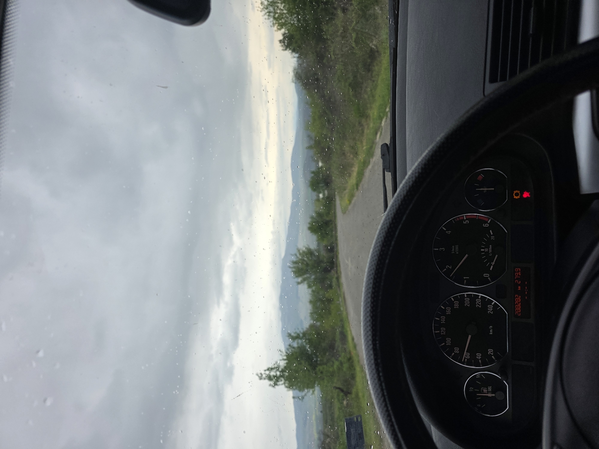 The image provides a driver's-seat perspective from inside a car, looking out through a rain-speckled windshield at a rural landscape. A winding asphalt road is visible through the windshield, extending into a valley surrounded by green, tree-covered hills and distant, hazy mountains under an overcast sky. Numerous raindrops are visible on the windshield, suggesting recent or ongoing precipitation.
The right side of the image features a portion of the car's interior, including the steering wheel and dashboard. The speedometer on the dashboard registers 0 km/h, indicating the vehicle is stationary or moving very slowly. The tachometer shows the engine idling at approximately 500 RPM (0.5 x1000 1/min). The odometer displays a total mileage of "288282 km", with a trip meter reading of "279.9 km". The fuel gauge indicates the tank is slightly above empty, and the word "DIESEL" confirms the fuel type. Several warning lights are illuminated on the dashboard, including a red general warning symbol (exclamation mark in a circle), an orange light that resembles an engine or system fault indicator, and another red light, possibly for brakes or another critical system.
The steering wheel, with a textured grip, is visible in the foreground. The weather is cloudy and appears to be daytime, with a subdued light cast over the mountainous or hilly terrain. The scene depicts a quiet, scenic road in a rural setting, consistent with the given location context of Lelintsi, Bulgaria.