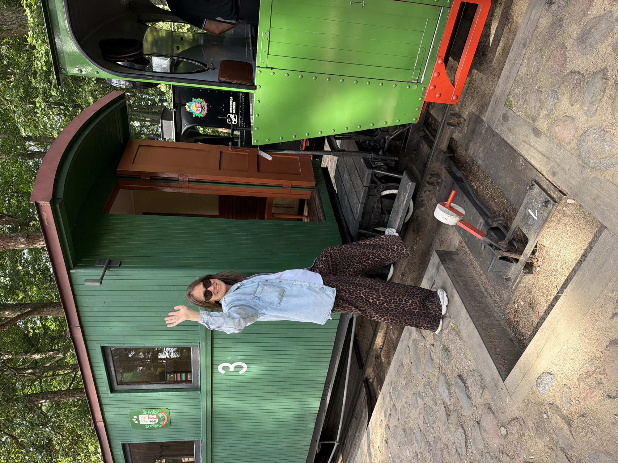 A woman wearing sunglasses, a denim jacket, and leopard print pants stands on a stone pathway next to a bright green train car. She is waving to the camera with her right hand, her left leg bent at the knee. The train car has a dark brown door ajar, revealing wooden benches inside. A smaller green train engine is visible to the right of the car. The scene appears to be set in an outdoor location with trees in the background. This is at the Ventspils Narrow Gauge Railway museum in Ventspils, Latvia.